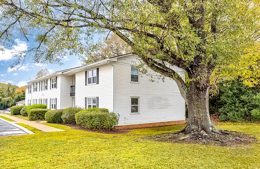 Charming white apartment complex surrounded by lush greenery and a large, majestic tree, offering tranquility and comfort in a serene neighborhood.