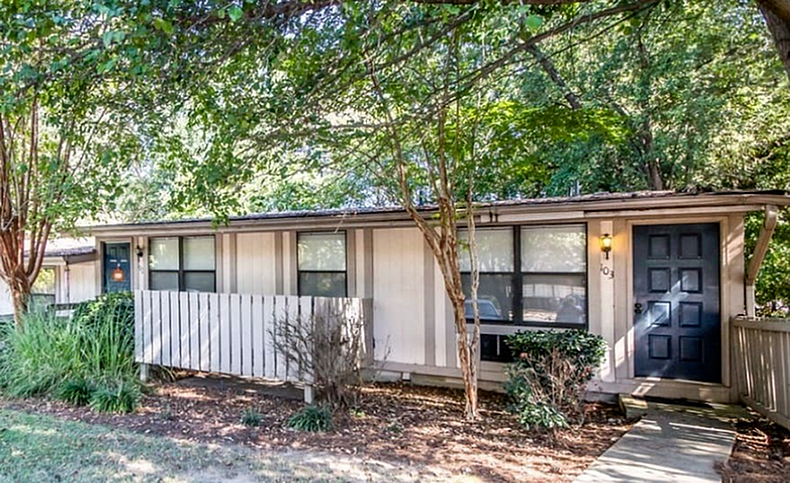 Charming single-story home with a modern black door, surrounded by lush greenery and inviting pathway.