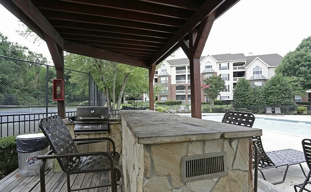 Charming outdoor area with a cozy gazebo, BBQ grill, and inviting seating, overlooking a serene pool and elegant apartment buildings.