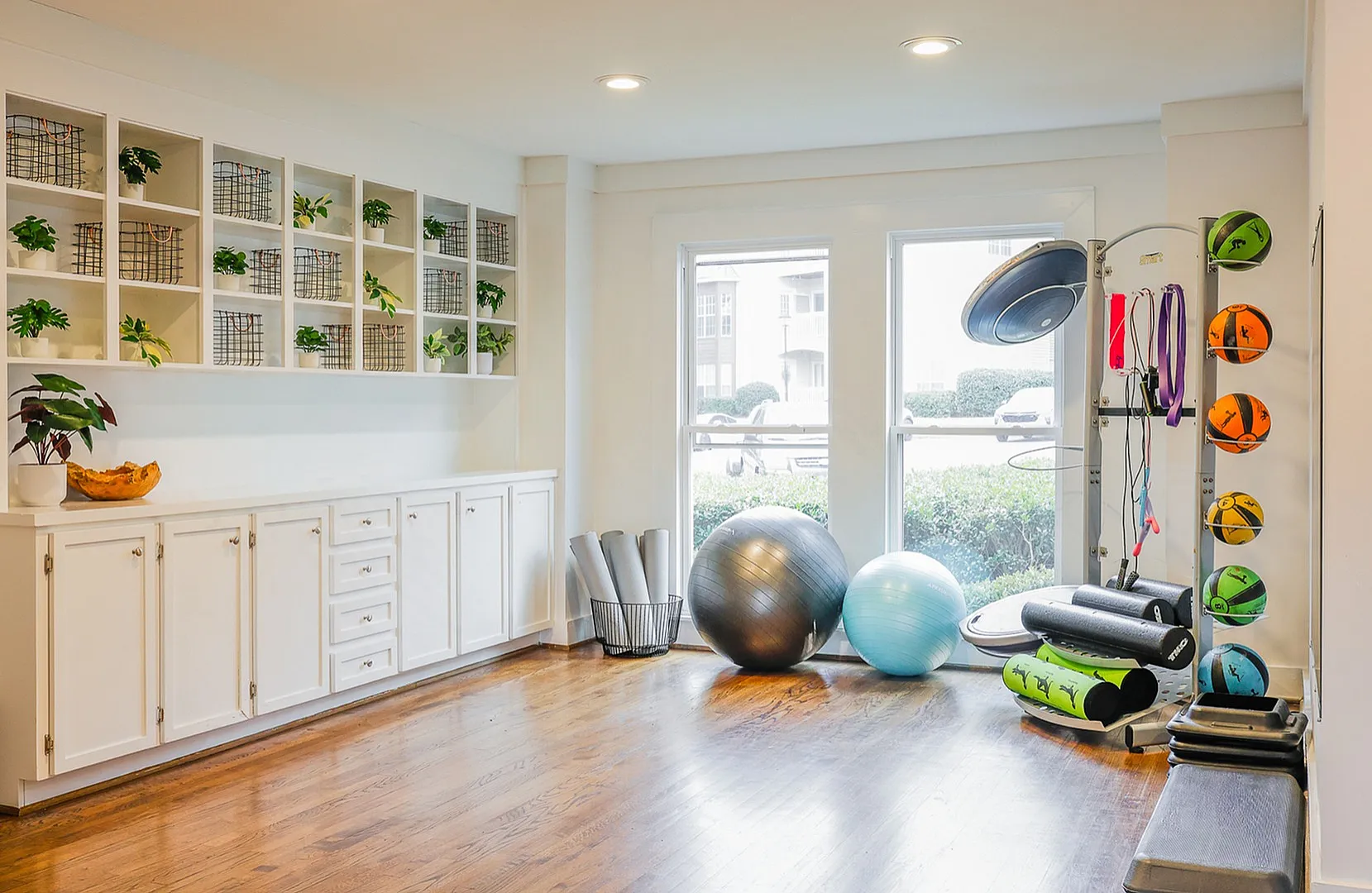 Inviting home gym with ample natural light, featuring vibrant exercise equipment, sleek white cabinetry, and a polished wood floor.