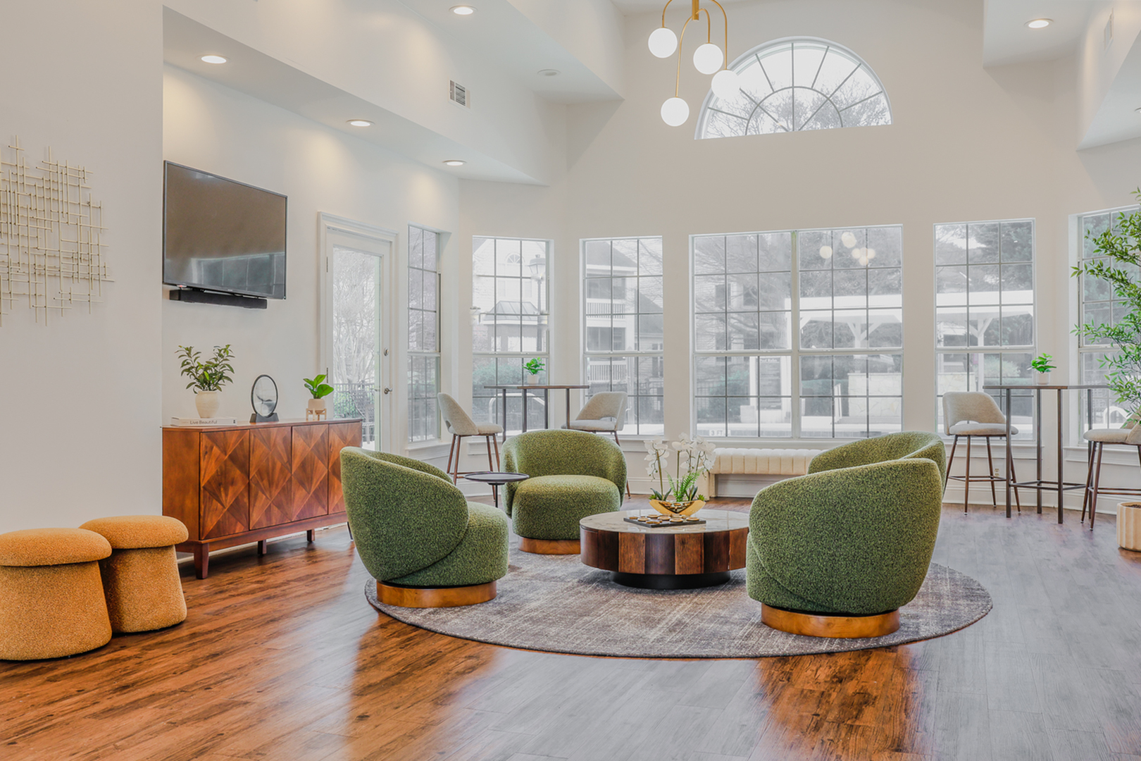 A warm and inviting clubhouse living space featuring chic green lounge chairs, an elegant round coffee table, and expansive sunlit windows that showcase serene views