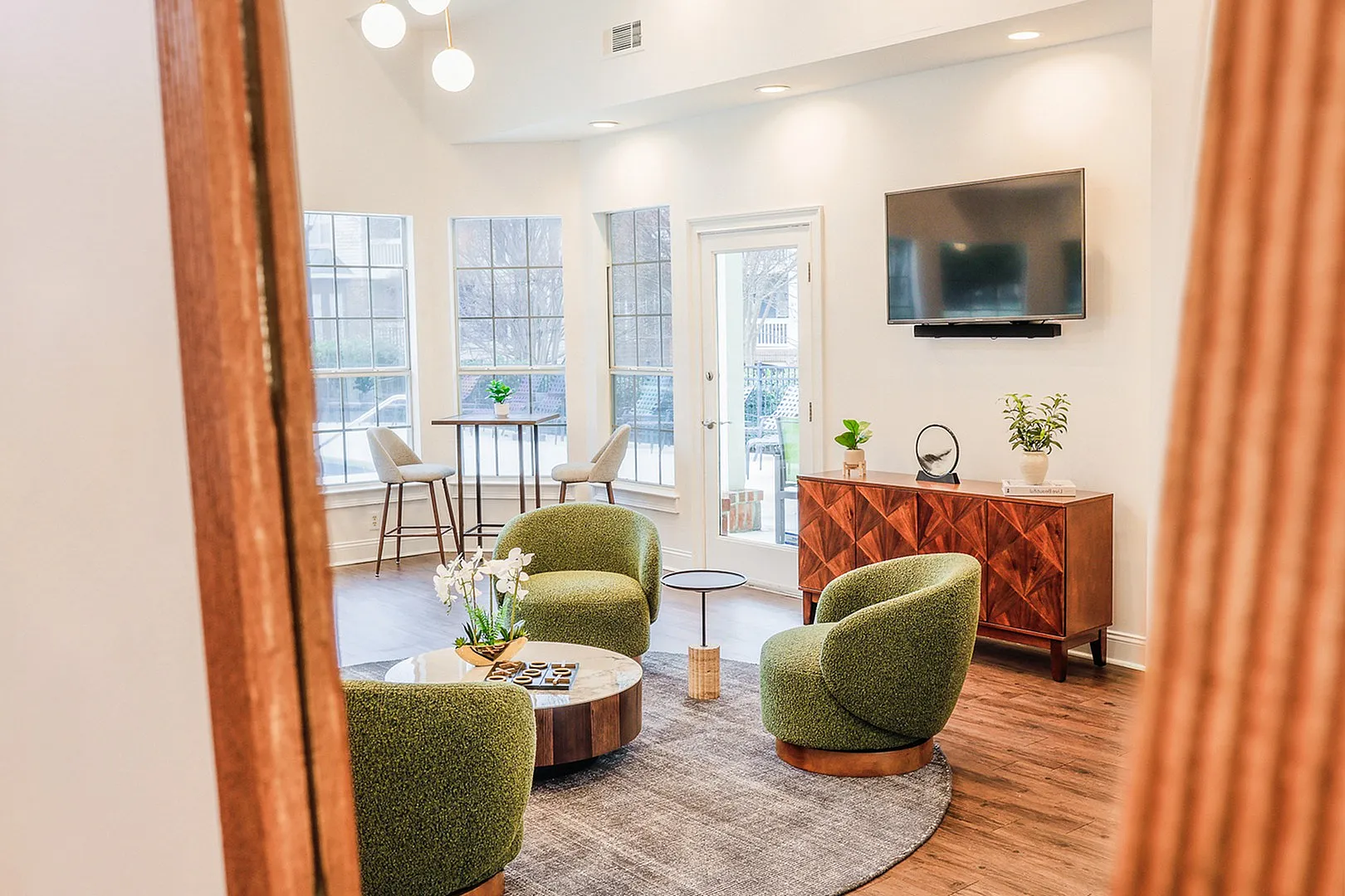 Bright and inviting clubhouse featuring modern green chairs, a stylish circular table, and sleek wood flooring. Expansive windows provide abundant natural light, while a chic media console and mounted TV complete the refined, contemporary atmosphere.