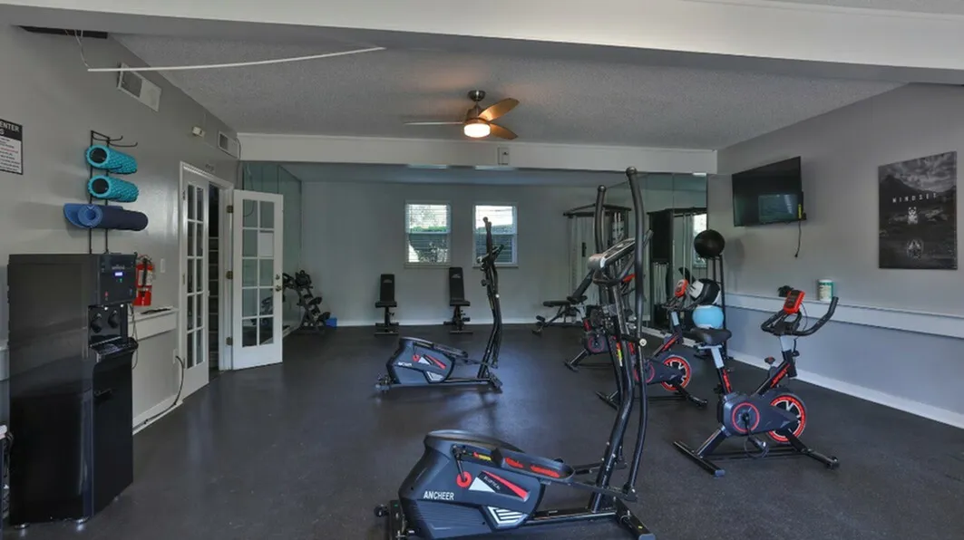 Modern fitness center with exercise bikes, free weights, and a wall-mounted TV in a spacious, well-lit room.