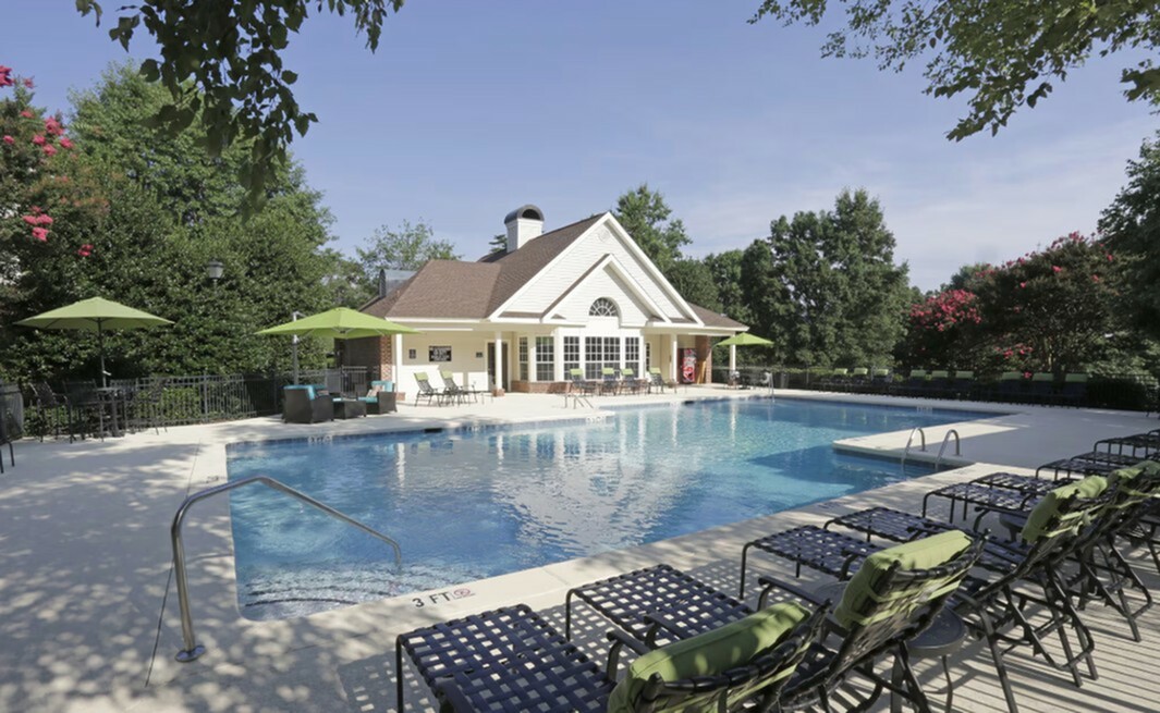 Inviting outdoor pool area with elegant loungers, umbrellas, and a charming clubhouse in a lush green setting.