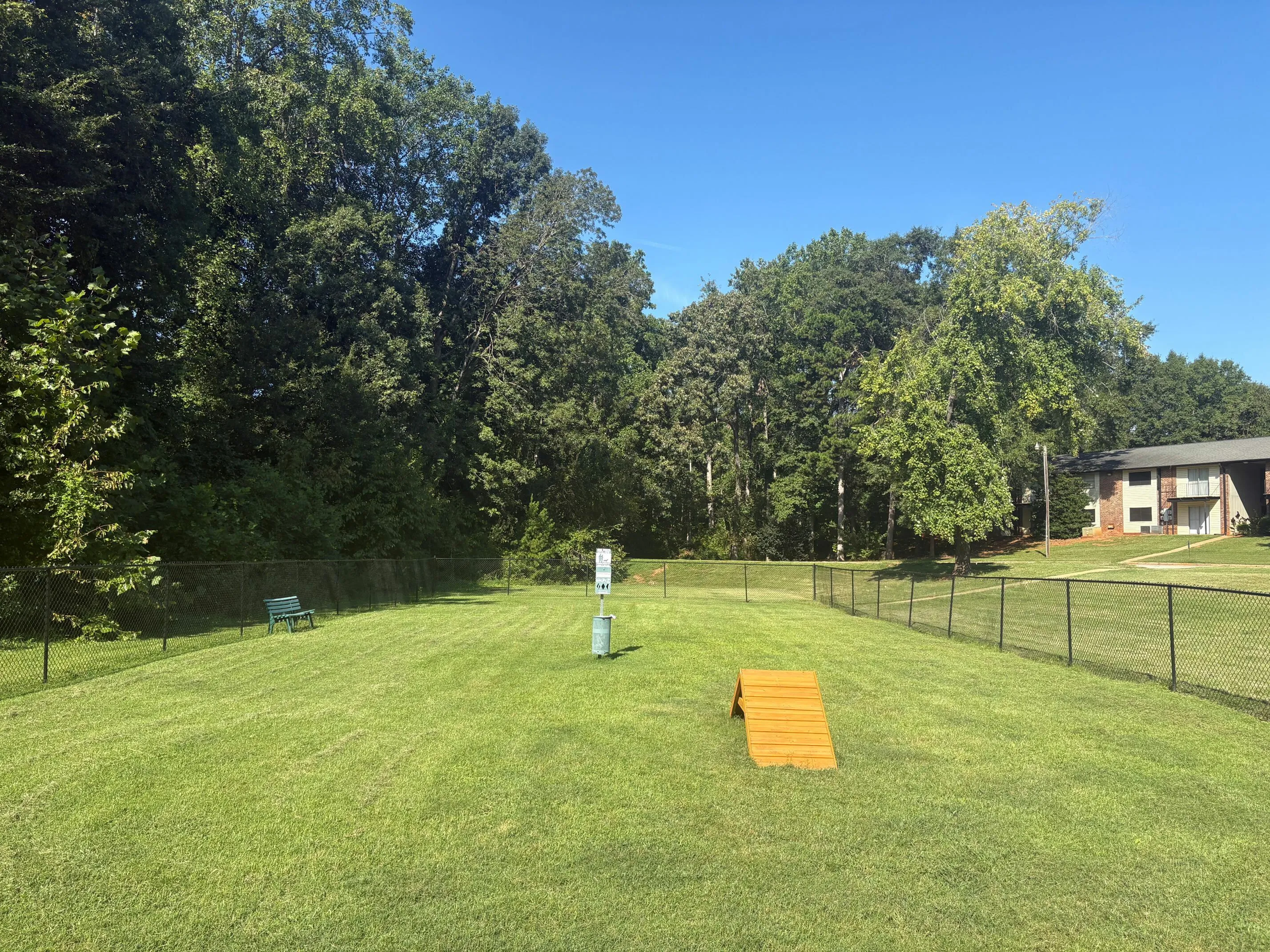 Expansive dog park featuring lush green grass, fenced area, a bench, and agility ramp, perfect for outdoor fun in a serene setting.