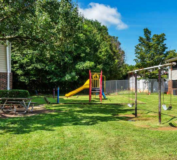 Charming community play area with vibrant slide, swings, and picnic tables surrounded by lush greenery.