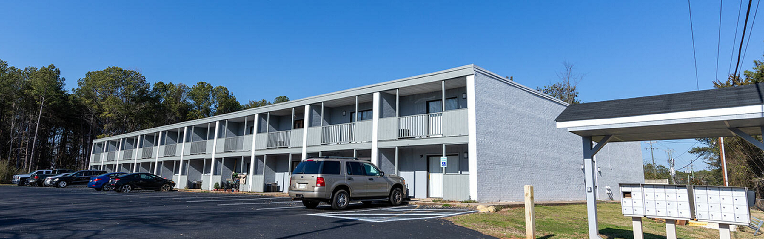 Modern two-story apartment complex with a spacious parking area and communal mailboxes, surrounded by lush greenery and blue skies.