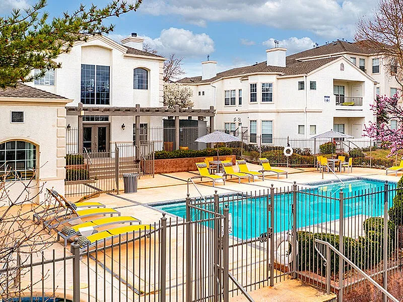 Charming apartment complex with a pristine pool, sun loungers, and vibrant outdoor seating, surrounded by elegant white buildings under a clear blue sky.