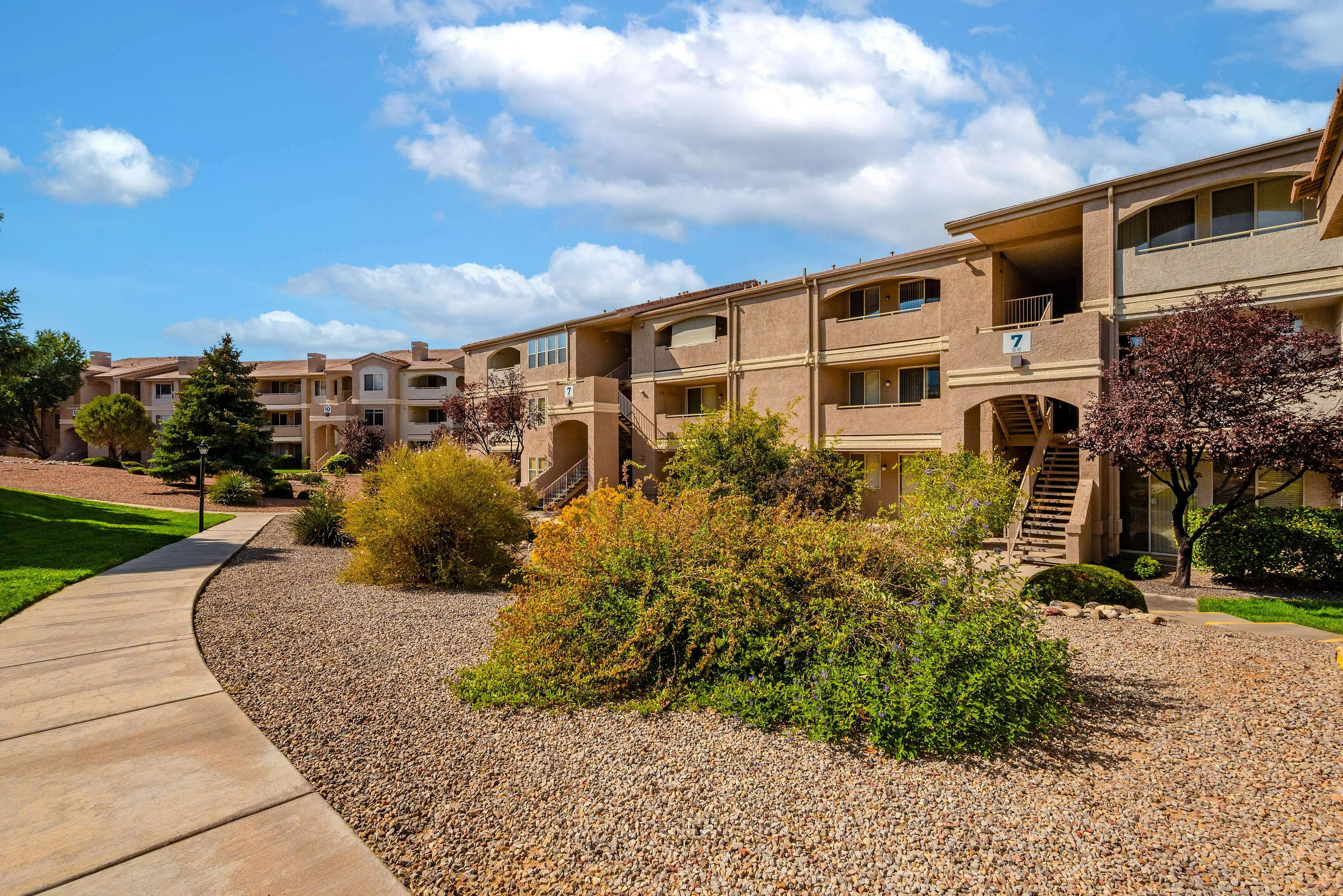 Charming apartment complex set amid lush greenery and landscaped pathways, offering a serene living environment under a bright blue sky.
