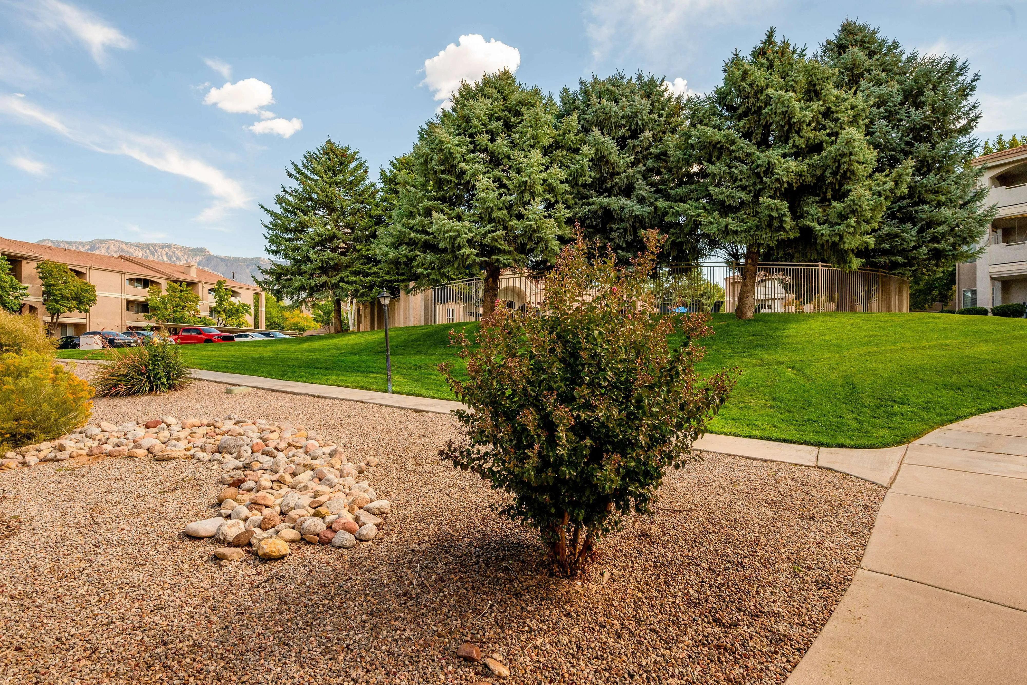 Serene apartment landscape showcasing lush green lawns, meticulously arranged gravel, and majestic trees with a backdrop of mountain views.