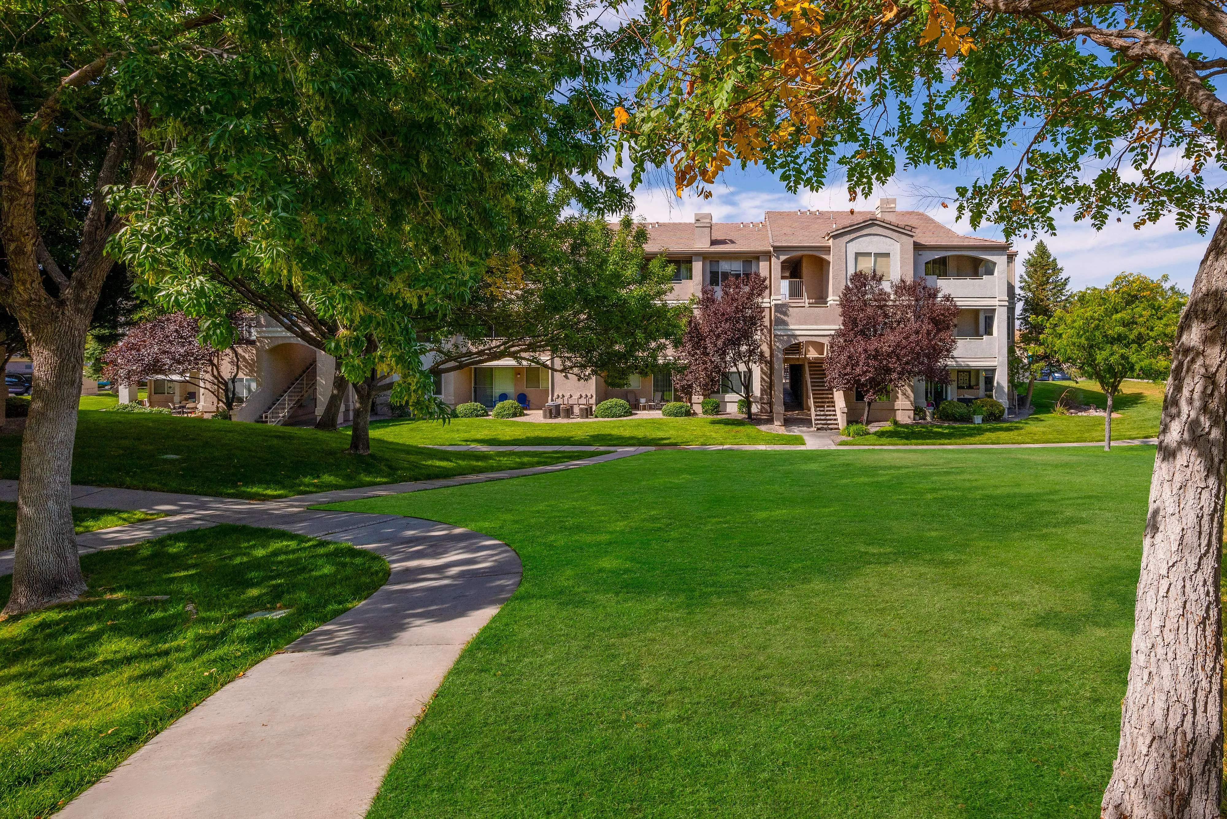 Inviting apartment view featuring lush green lawns, mature trees, and elegant multi-story architecture under a clear blue sky.