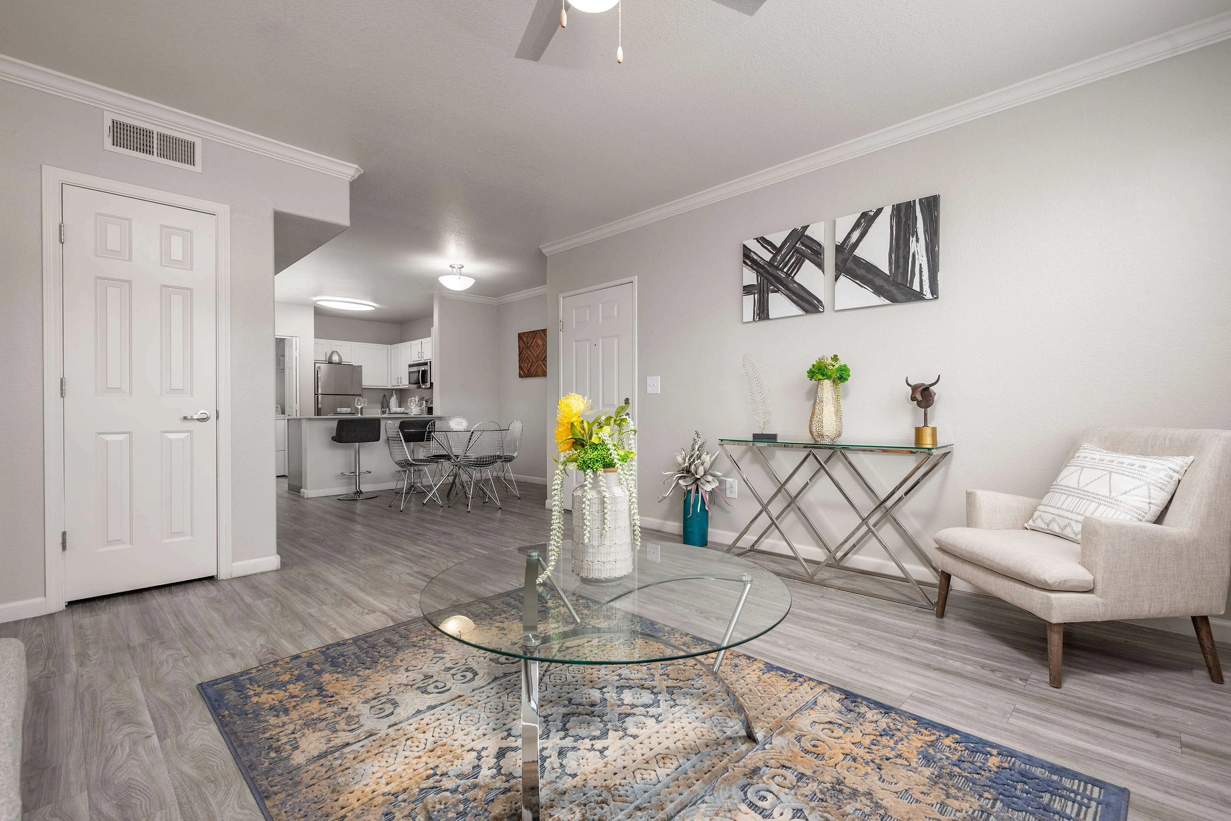 Bright and modern living area with elegant decor and open-concept design, featuring a stylish glass coffee table, cozy seating, and sleek kitchen in the background.