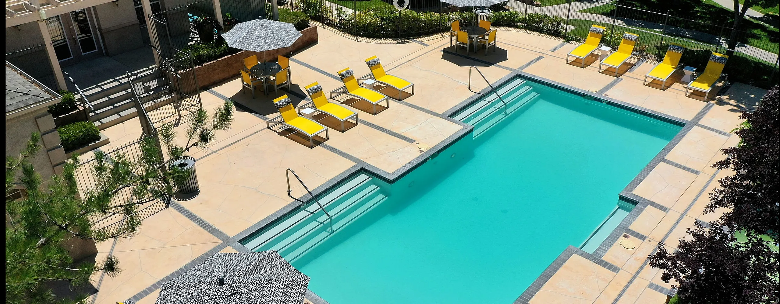 Luxurious apartment complex pool area with inviting turquoise waters, surrounded by vibrant yellow sun loungers and stylish patio umbrellas, complemented by lush greenery.
