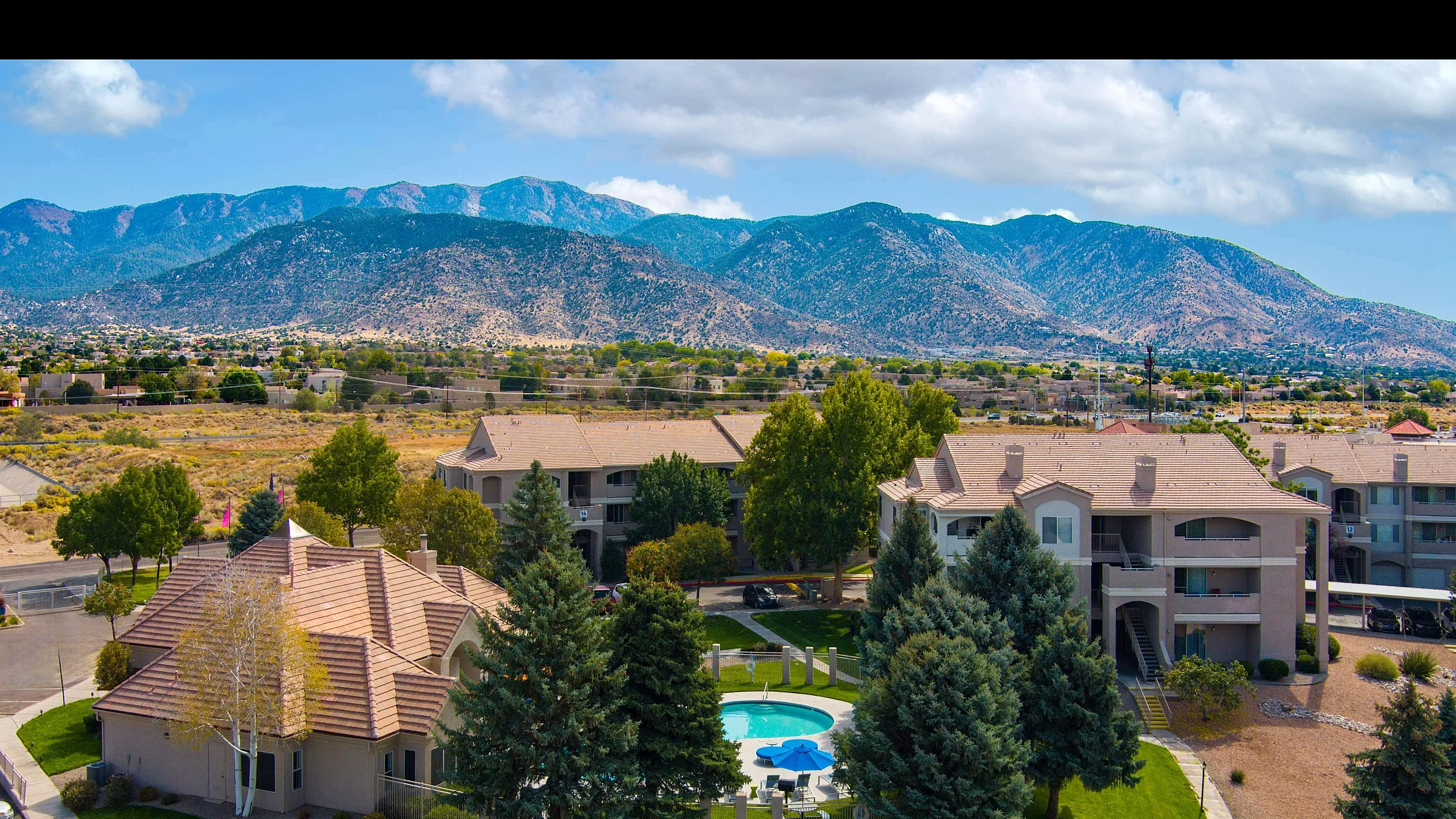 Charming apartment complex with lush green landscaping, a sparkling central pool, shaded seating, and scenic mountain views.