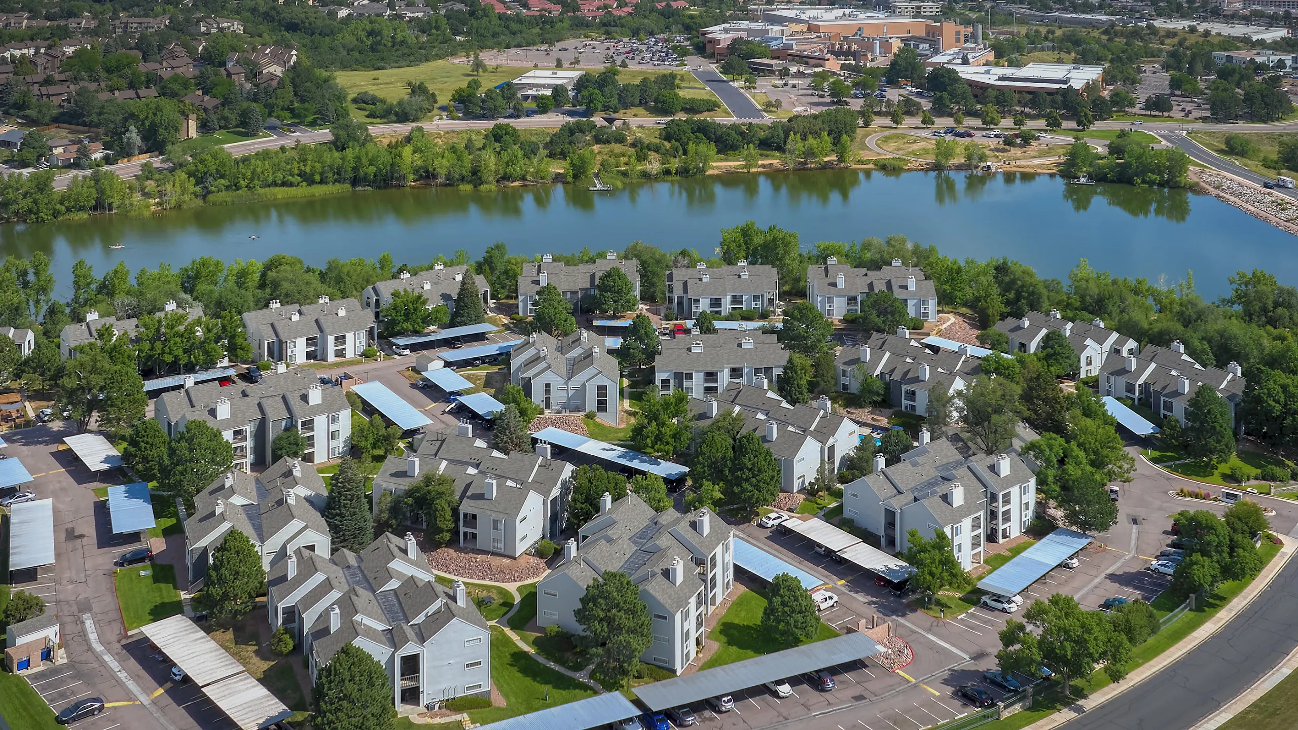 Scenic aerial view of modern apartment complex with lush greenery and a tranquil lake setting, offering desirable living in a vibrant community.