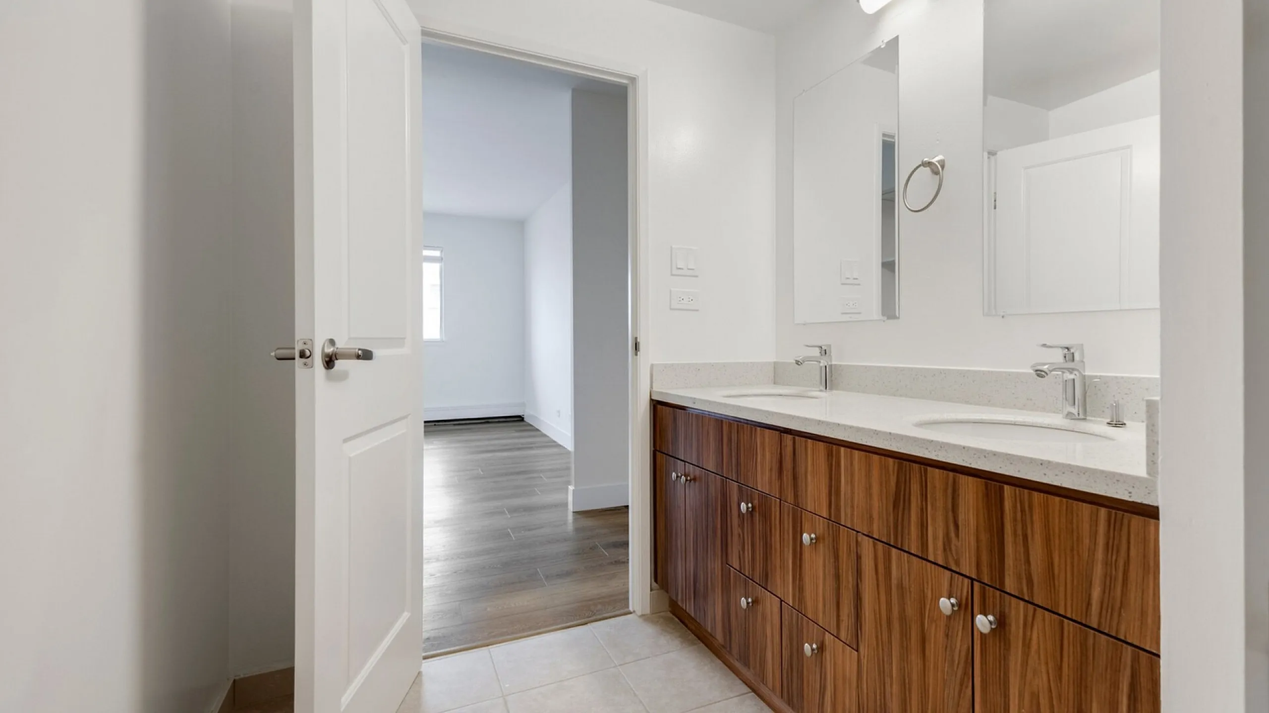 Elegant bathroom with dual sinks, wood cabinetry, and a bright doorway leading to a spacious, light-filled room with sleek wood flooring.
