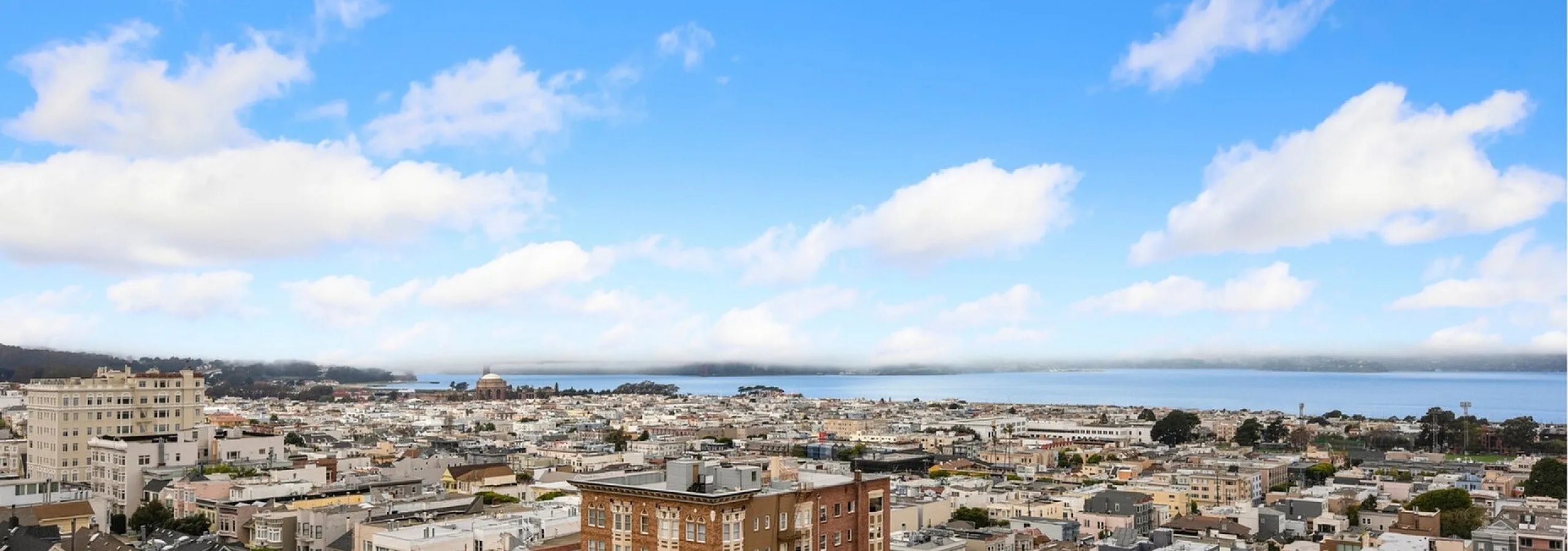 Breathtaking panoramic view of a vibrant cityscape, overlooking a tranquil bay under clear blue skies and fluffy clouds.