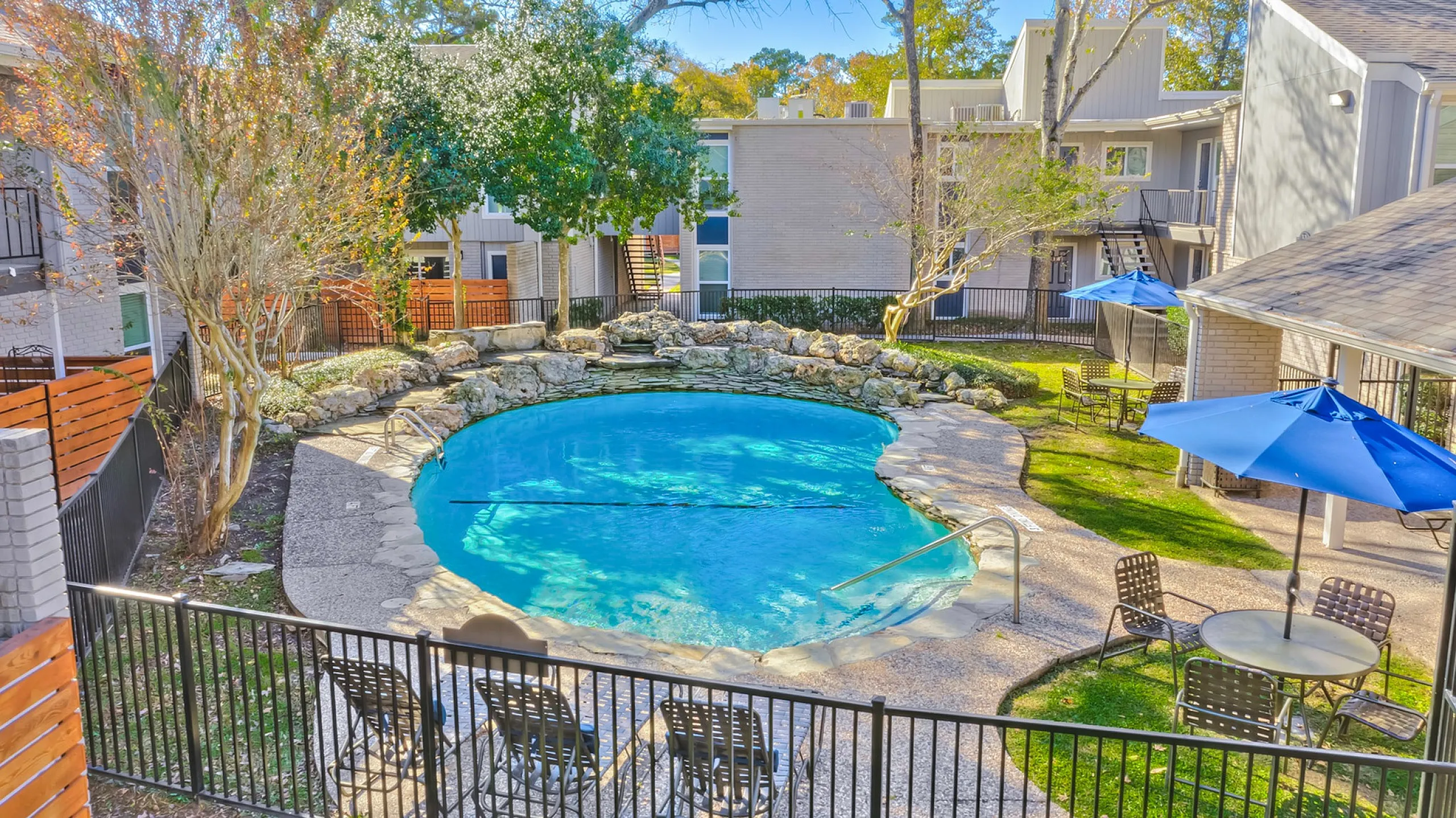 Inviting apartment complex pool area with serene landscaping and vibrant blue umbrellas, perfect for relaxation and social gatherings.