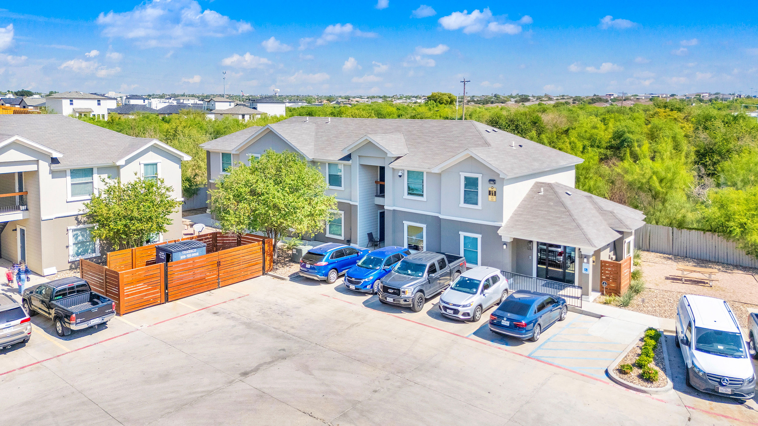 Modern apartment complex with ample parking and lush greenery, showcasing a bright and welcoming facade under a clear blue sky.