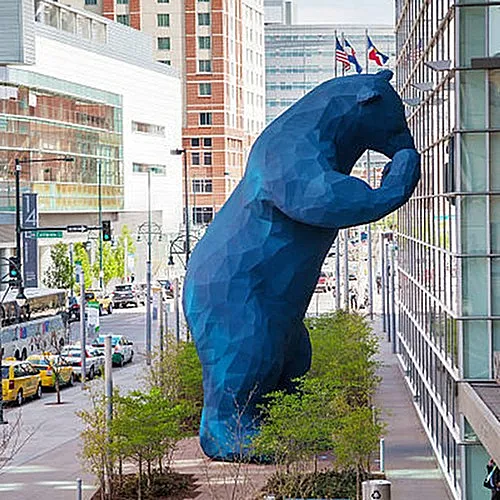 Vibrant city scene featuring a giant blue bear sculpture playfully peering into a building, surrounded by greenery and urban architecture.