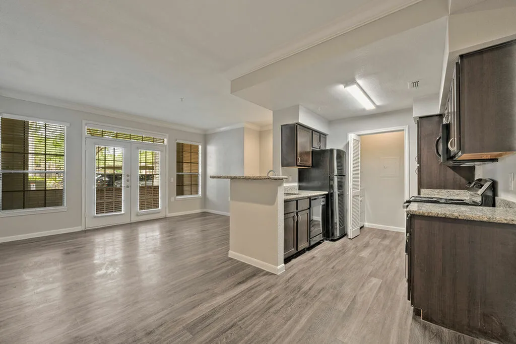 Modern open-concept living area with sleek dark cabinetry, spacious kitchen island, and expansive windows. Abundant natural light and elegant flooring complete this stylish apartment layout.