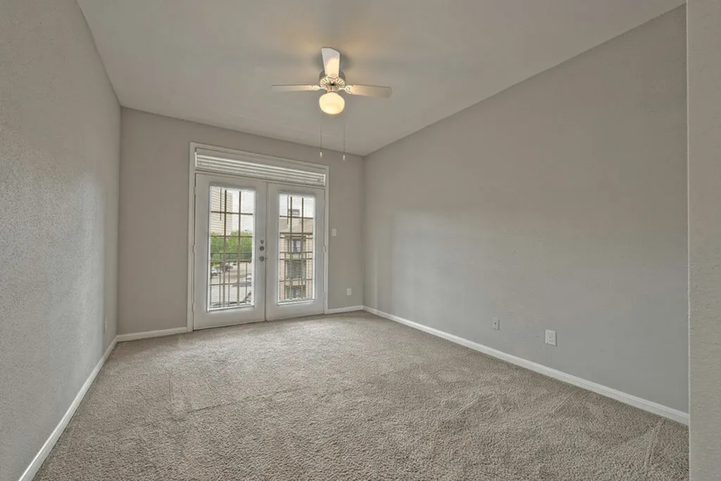 Bright apartment bedroom with soft carpeting, ceiling fan, and French doors leading to a balcony.