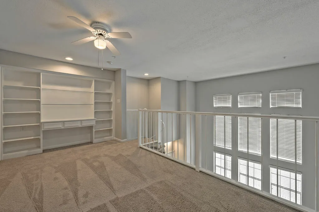 Bright open loft area with built-in shelving, ceiling fan, and large windows for natural light.