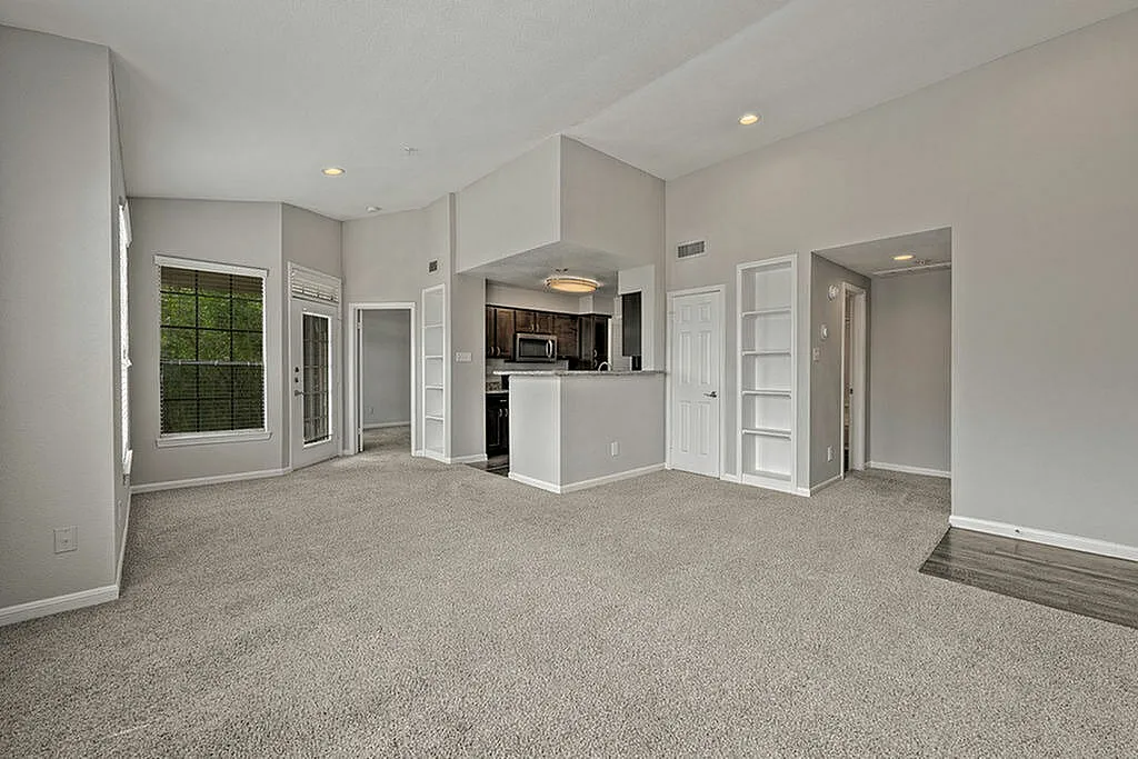 Spacious living area with modern open plan design, featuring plush carpet, built-in shelving, and a view into a sleek kitchen.