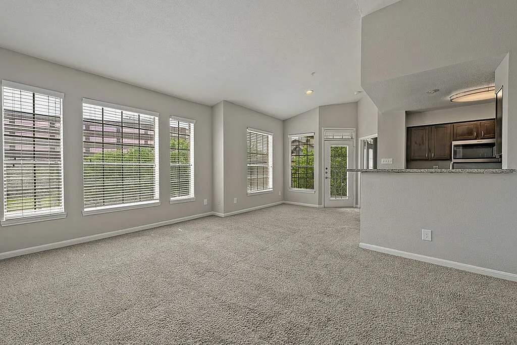 Bright and spacious living area with large windows, plush carpet, and an open-concept kitchen featuring modern cabinetry and granite countertops.