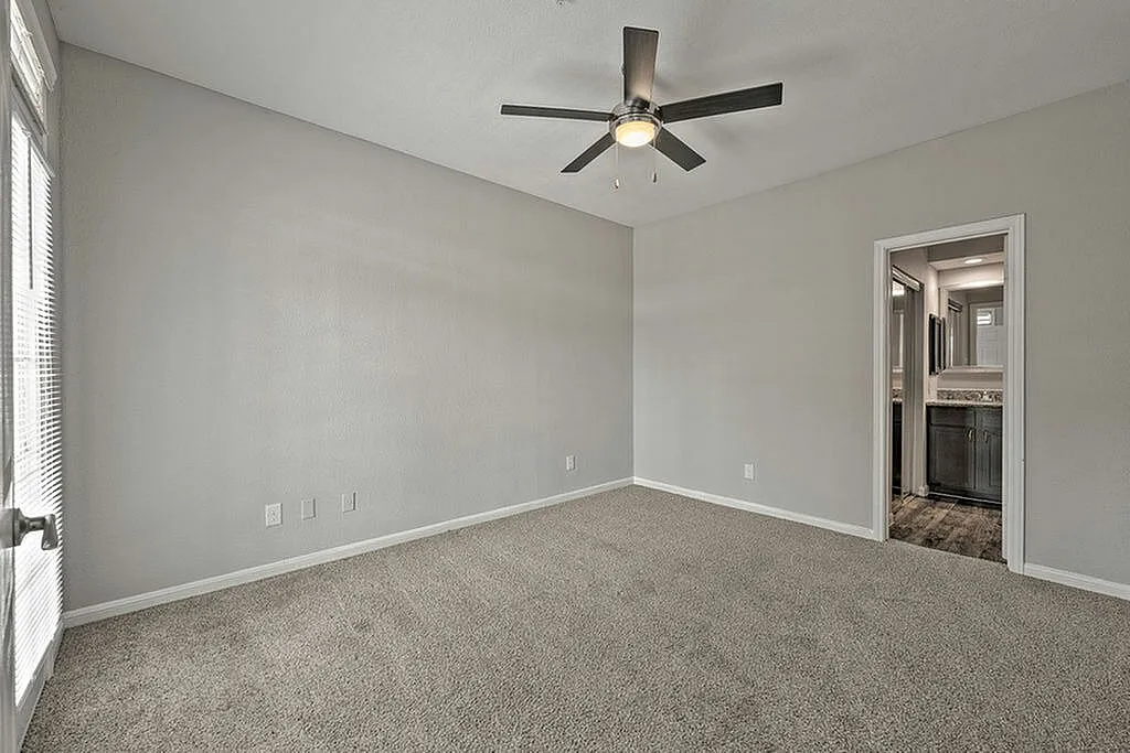 Spacious bedroom with plush carpeting, ceiling fan, and connected modern bathroom, filled with natural light from large windows.