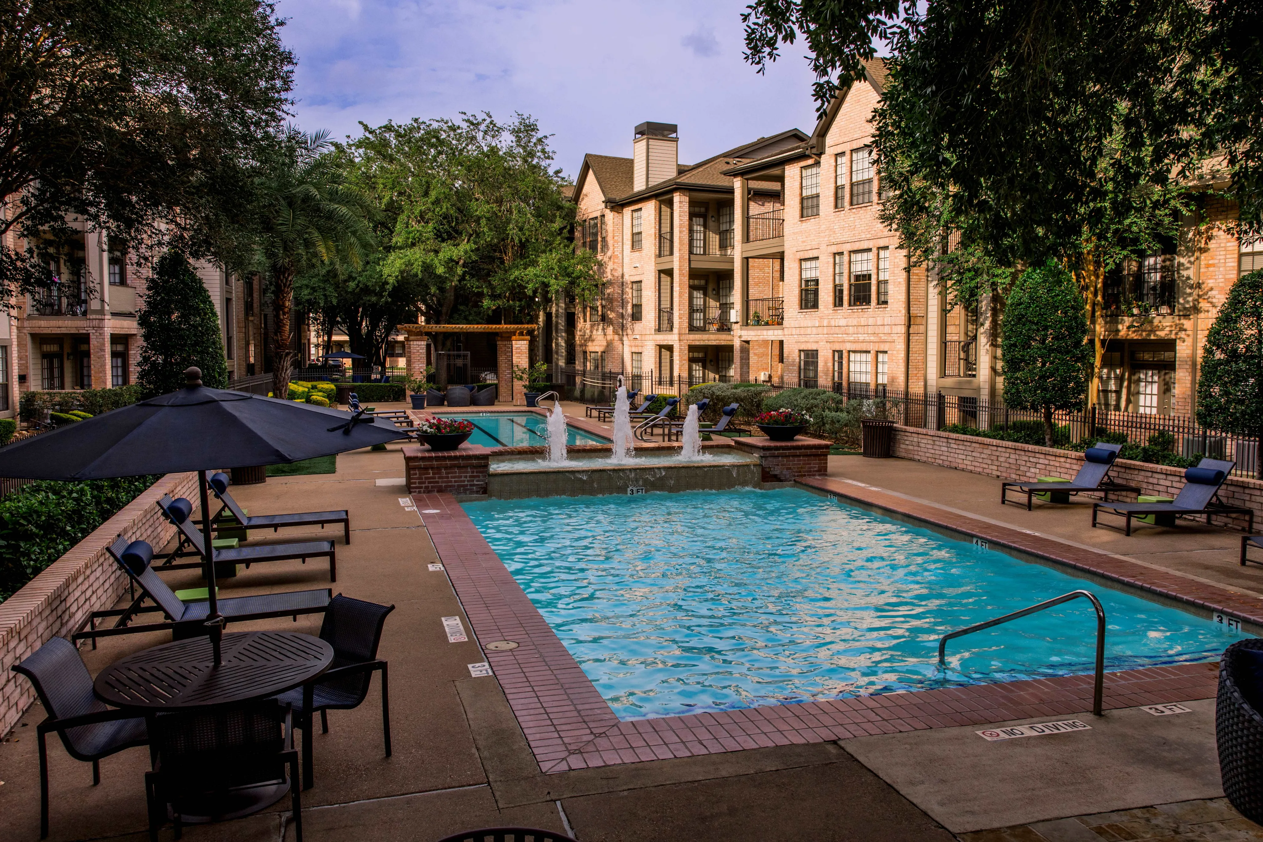 Charming apartment complex with a serene swimming pool, surrounded by lush greenery and elegant brick buildings.