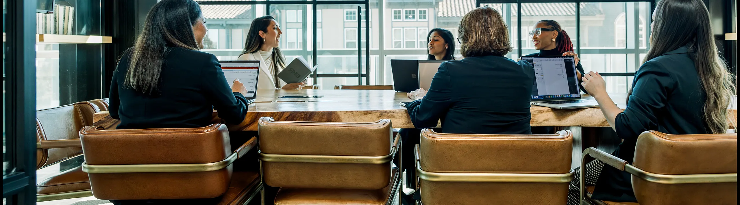 Modern conference room with large windows featuring a vibrant business meeting around a sleek wooden table with stylish leather chairs.