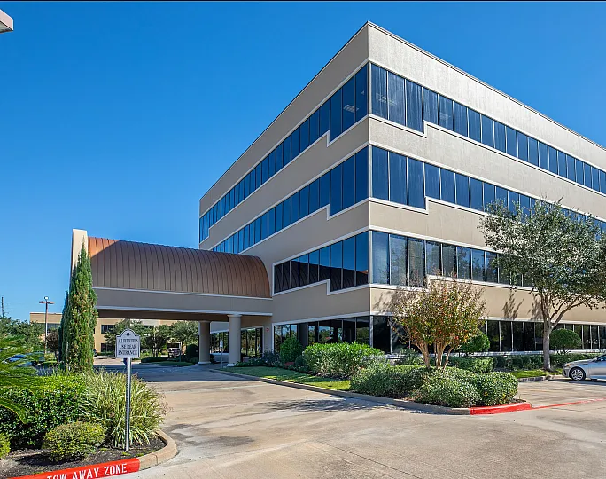 Modern office building with sleek glass windows, lush landscaping, and ample parking space under a clear blue sky.