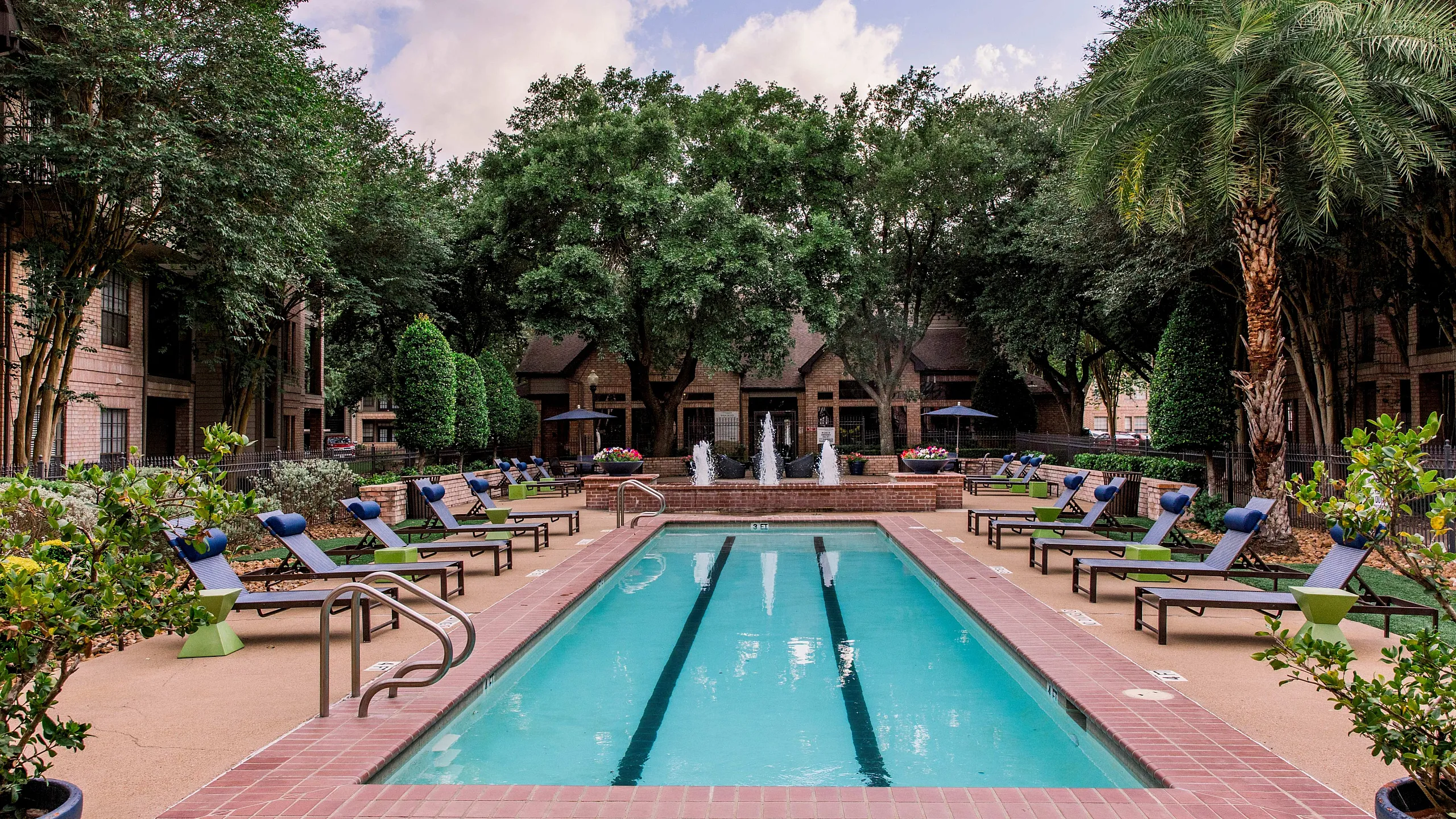 Elegant apartment pool area with luxurious lounge chairs, surrounded by lush greenery and scenic fountains.