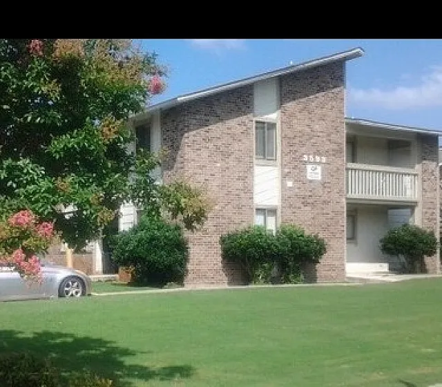 Charming apartment complex with lush greenery, vibrant flowers, and inviting brick exterior on a sunny day.