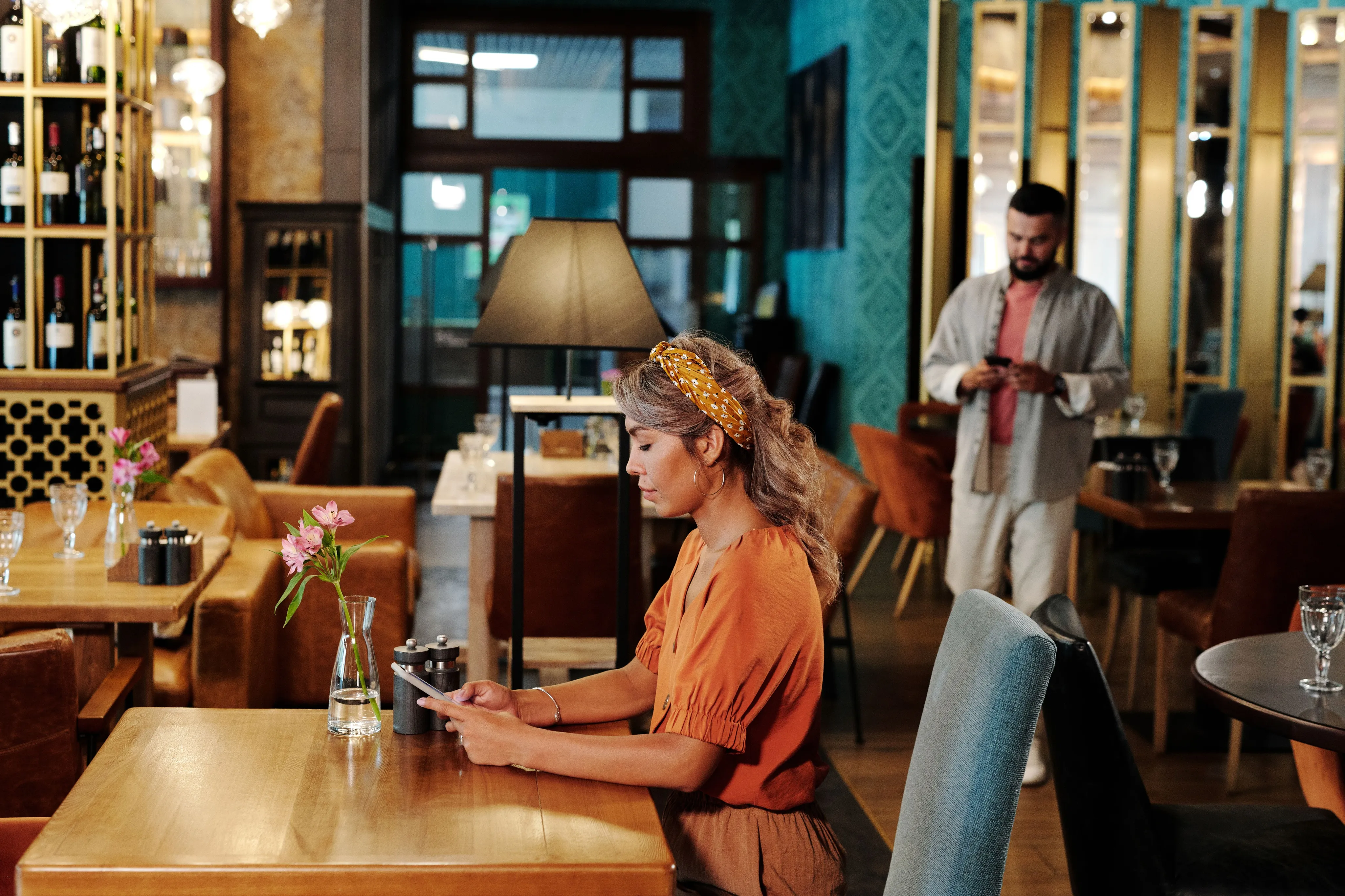 A woman wearing an orange shirt and yellow headband sitting at a wooden table using a smartphone inside a restaurant.