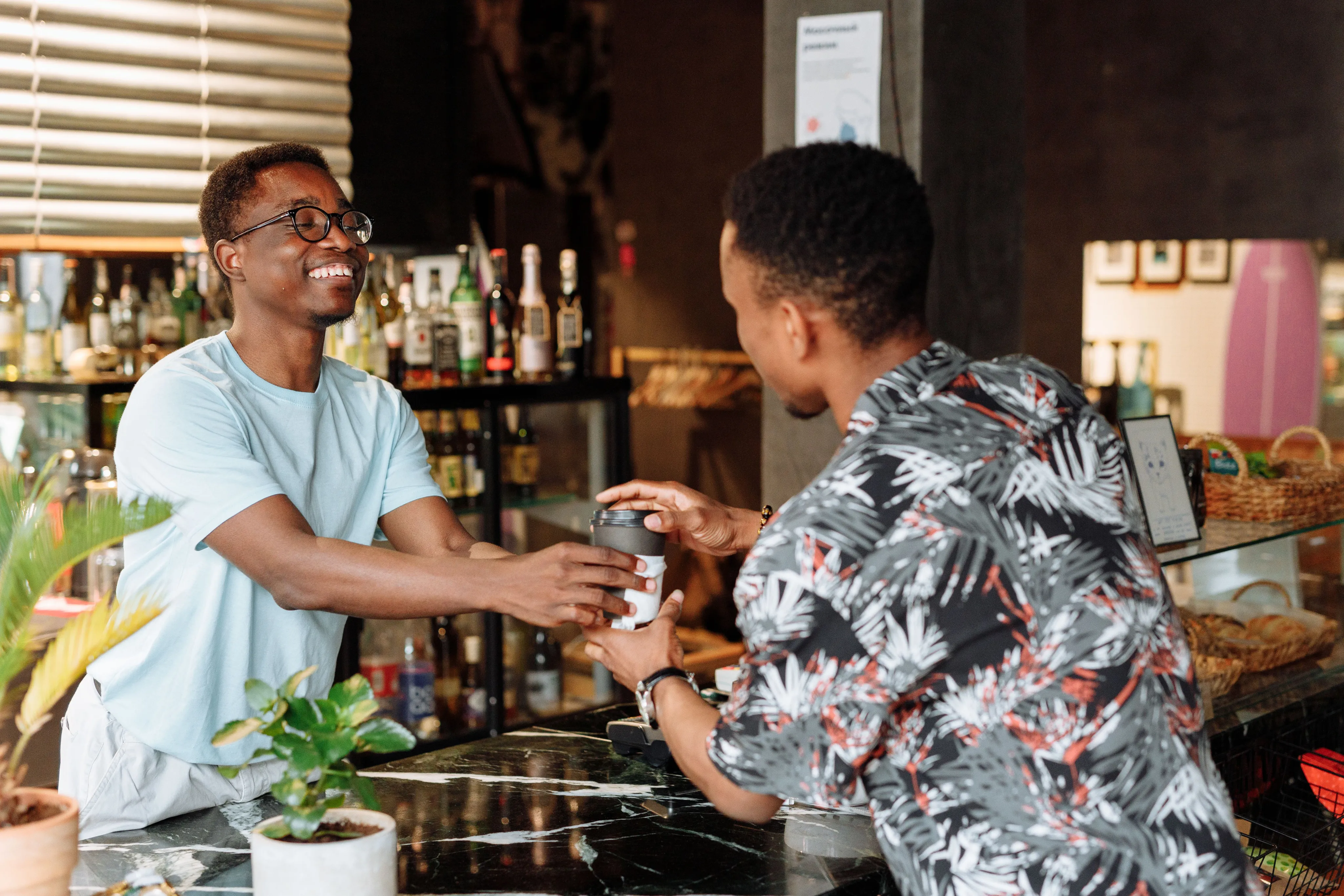 A smiling barista wearing glasses handing a paper coffee cup to a customer standing at a marble counter in a cafe.