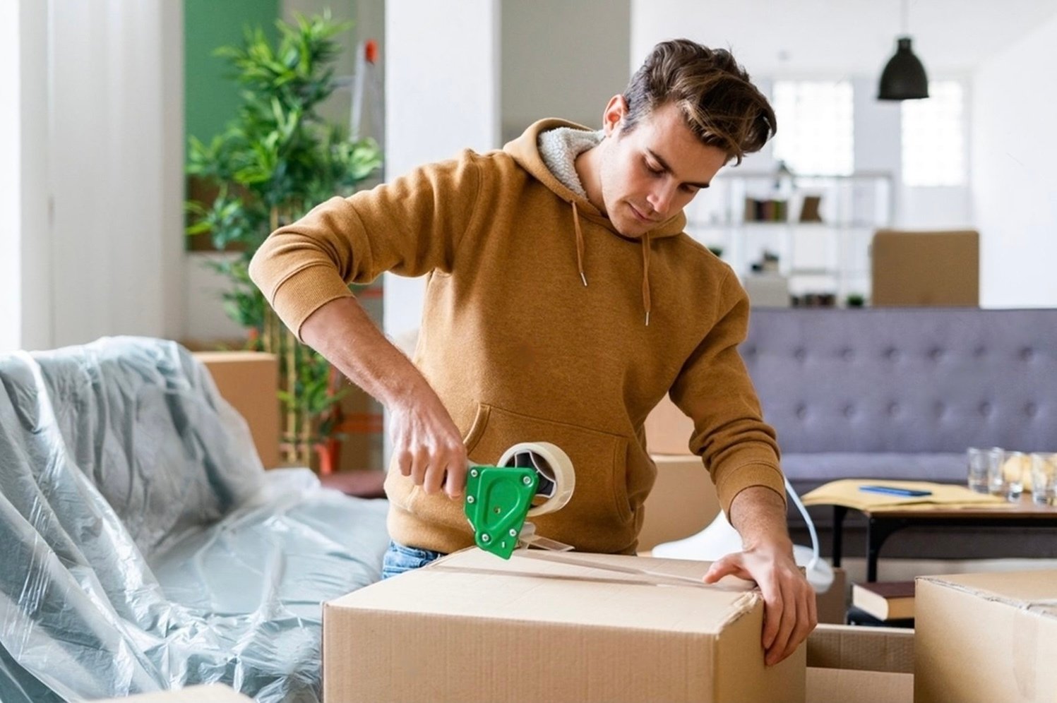 A man in a brown hoodie tapes a cardboard box in a living room filled with packed items and a covered sofa as he prepares for his move into a new Downtown Sarasota apartment at The DeSOTA.