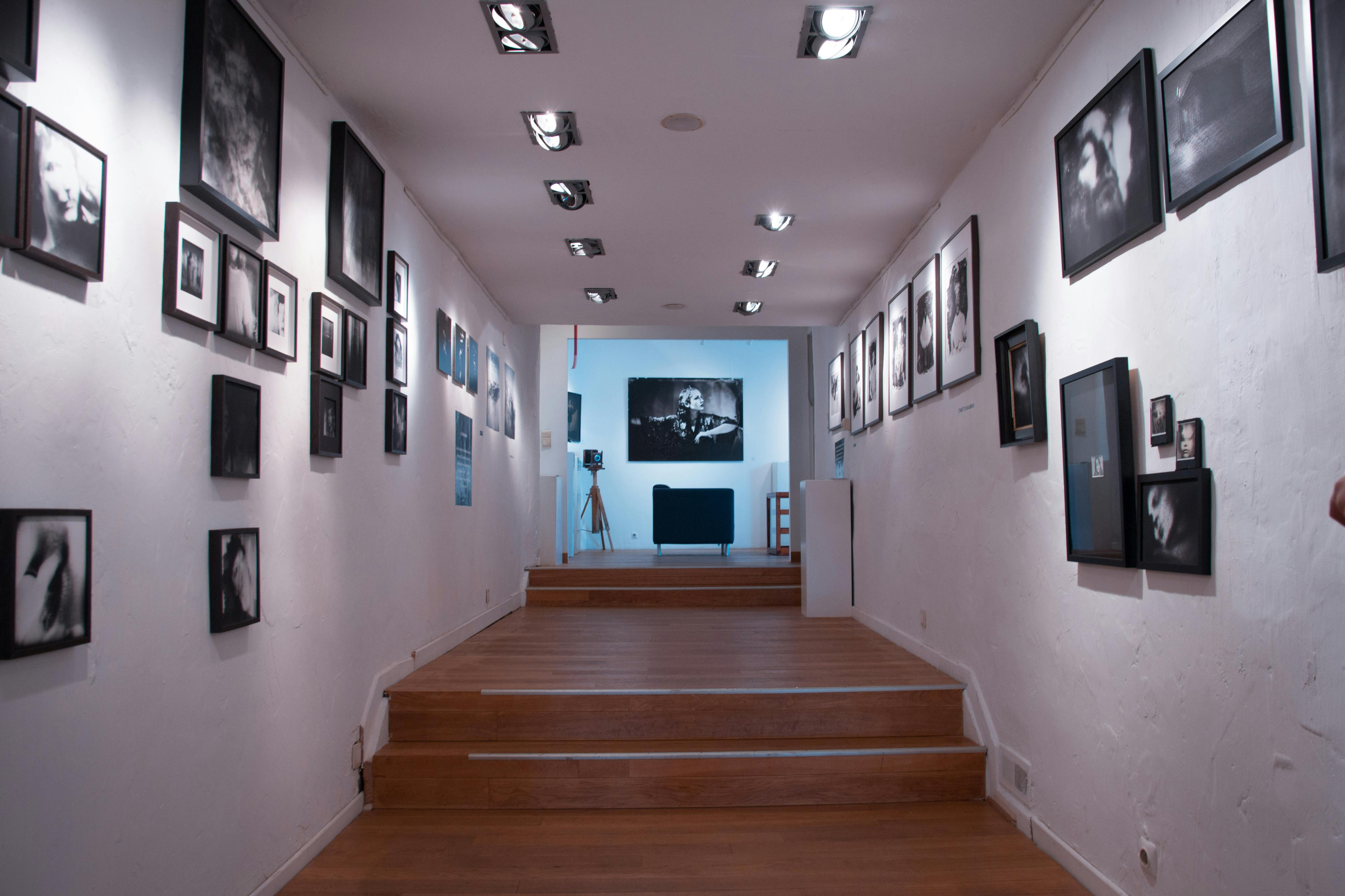 Black framed photos hanging on white walls along a wooden hallway featuring short stairs leading toward a lit room.