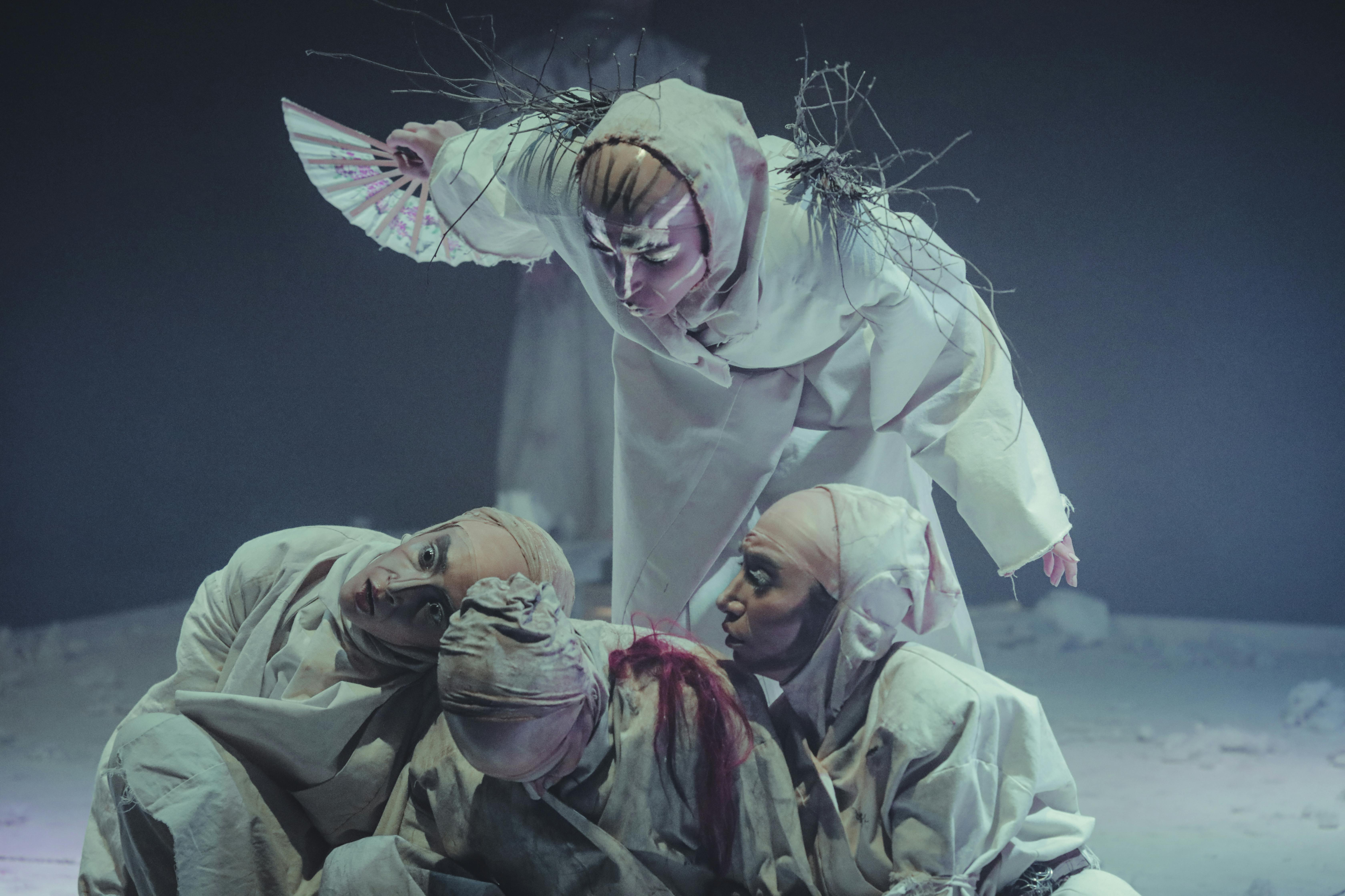 A performer wearing white clothing and twigs holding a fan leaning over three people crouching in white fabric.