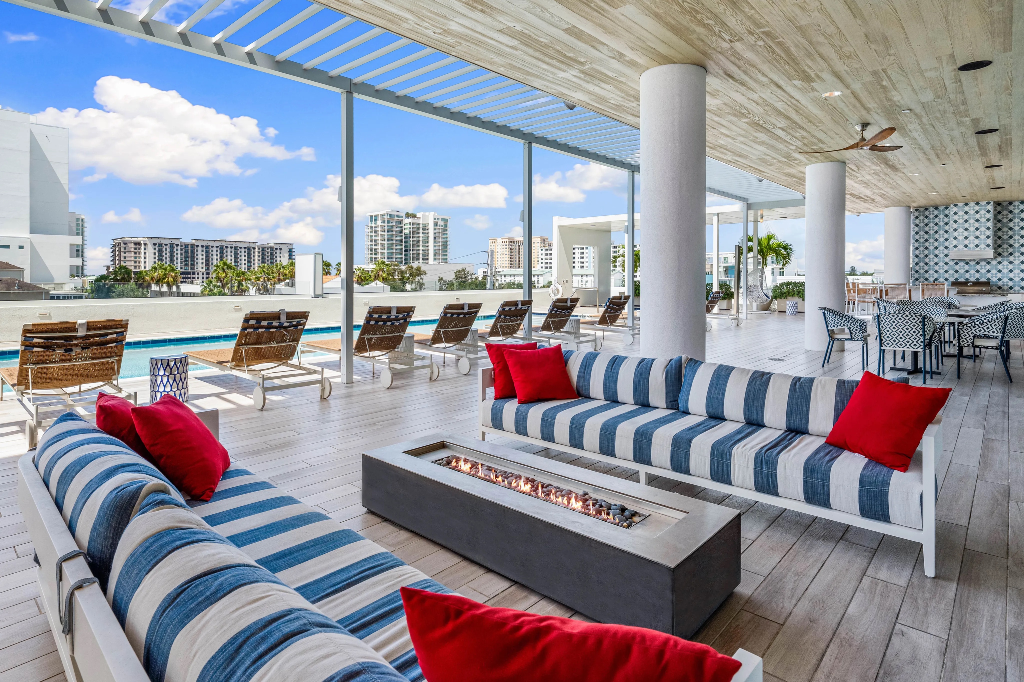Blue and white striped sofas displaying red pillows resting near a long fire pit and a swimming pool under a covered patio.
