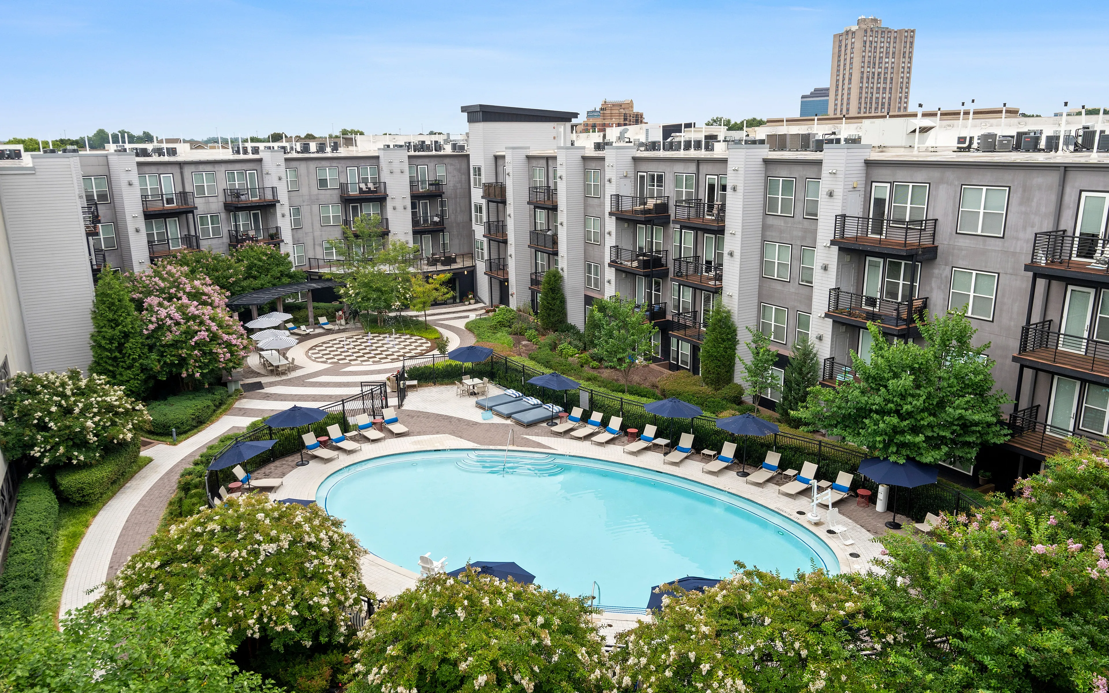 Modern apartment complex featuring a large oval swimming pool surrounded by lounge chairs and lush greenery.
