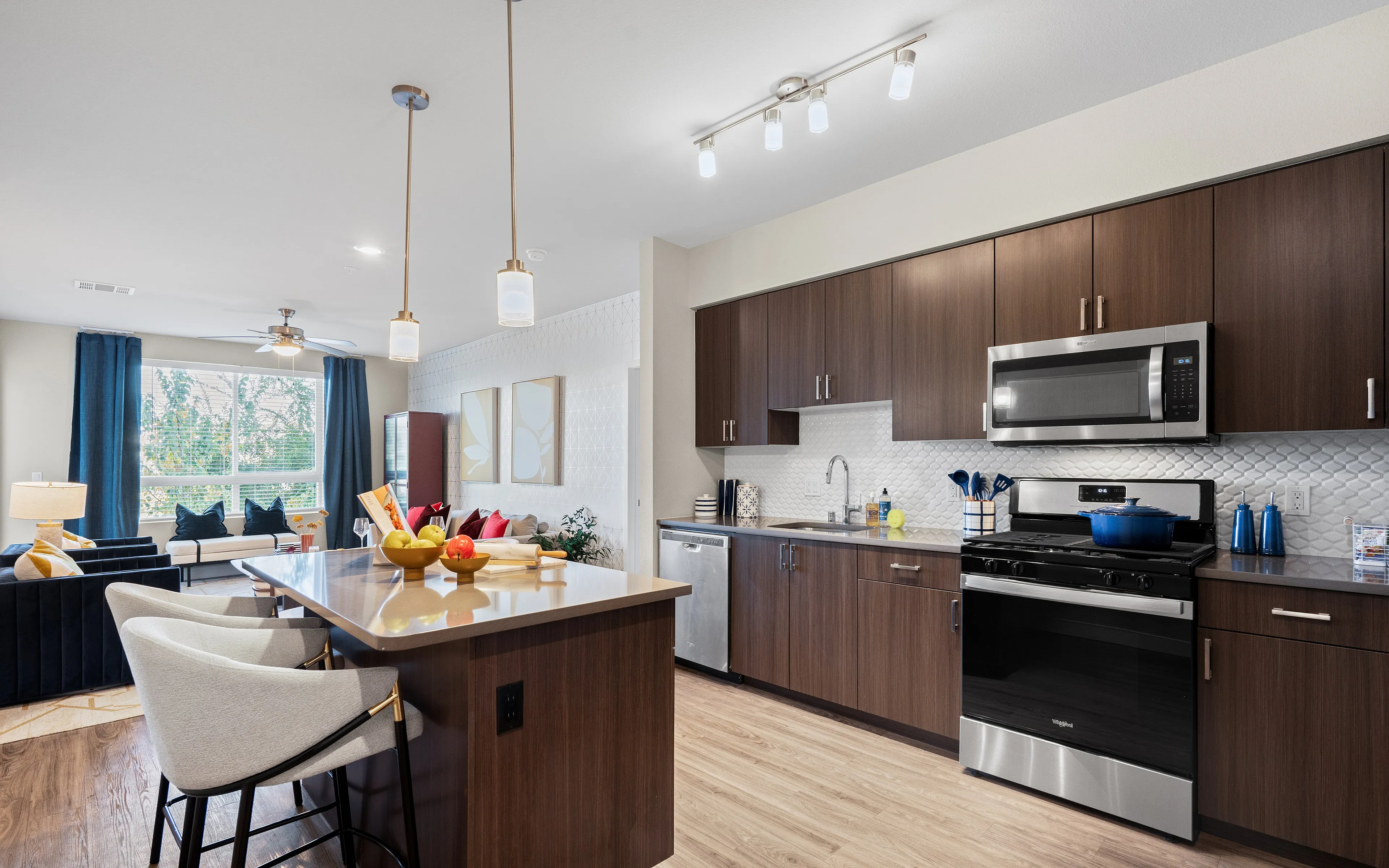 Contemporary open-plan kitchen and living room with elegant decor, featuring dark wood cabinetry, stainless steel appliances, large central island with bar seating, and modern lighting. Natural light floods the space through large windows w
