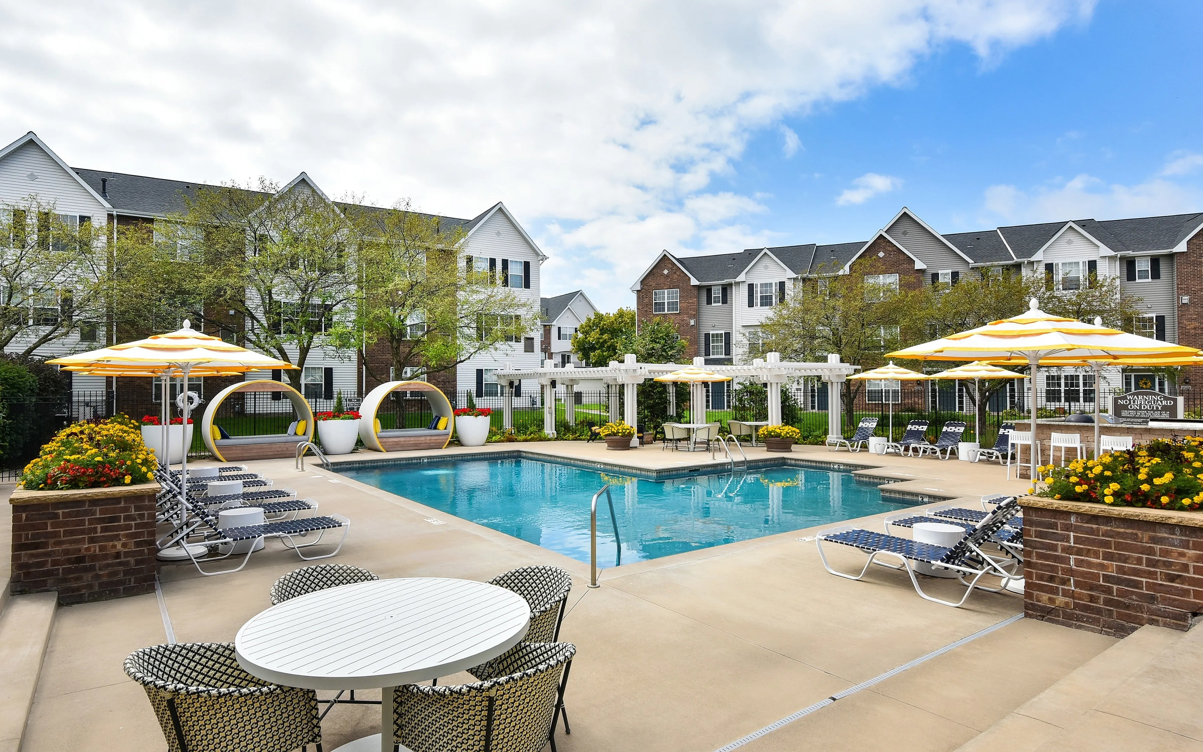 Charming apartment complex boasting a vibrant pool area with stylish lounge chairs and yellow umbrellas, surrounded by lush greenery and elegant buildings.