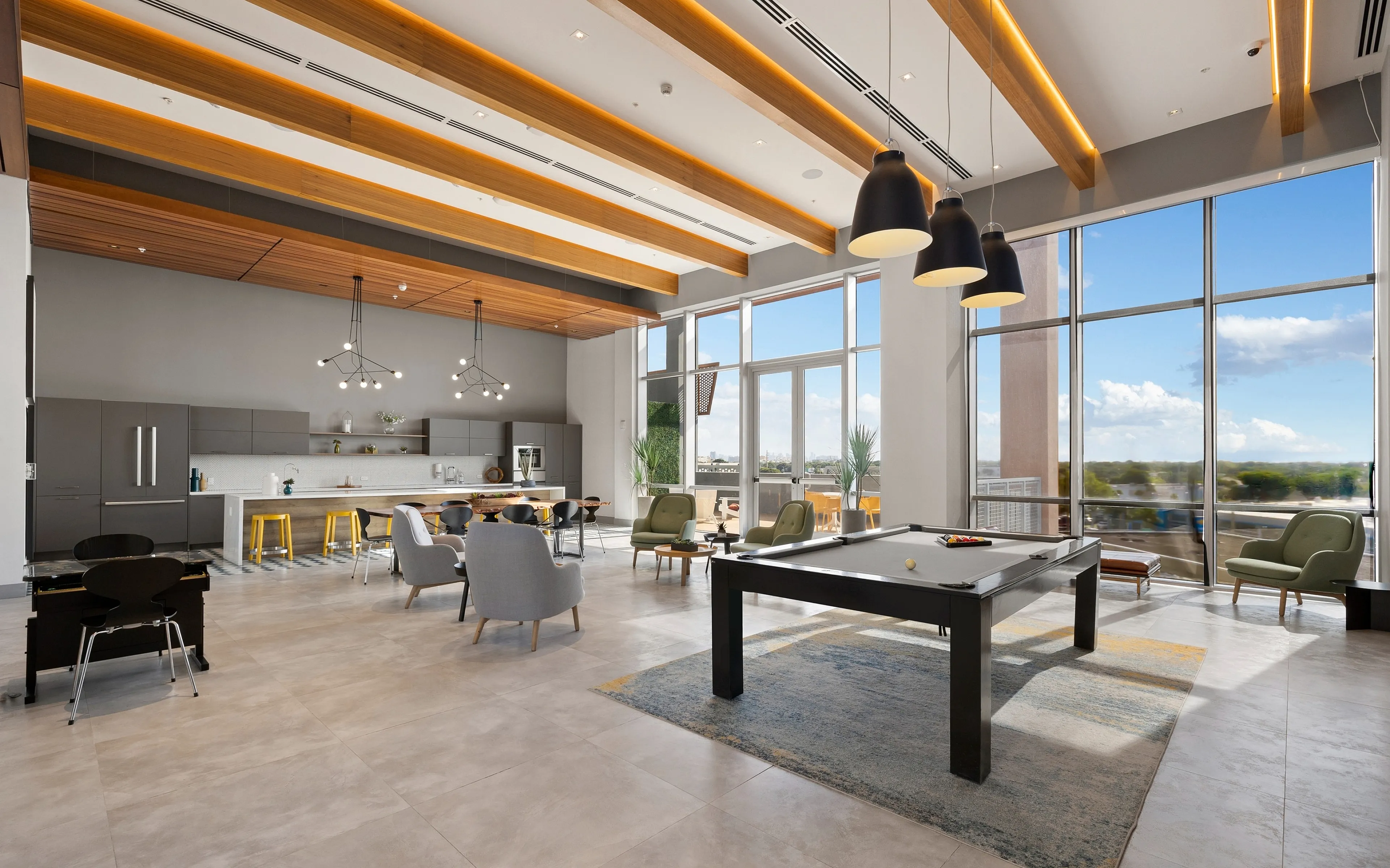Luxurious open-concept living area featuring a modern kitchen, high ceilings with wooden beams, and a stylish billiard table, all bathed in natural light from expansive windows.