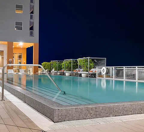 Luxurious rooftop pool area with sleek lounge chairs, vibrant greenery, and modern lighting against a clear night sky.