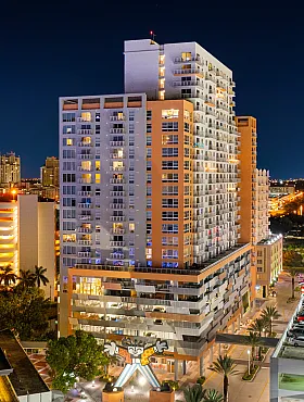 Stunning modern high-rise apartment building illuminated at night, surrounded by vibrant city lights and palm trees, showcasing urban luxury living with convenient parking.
