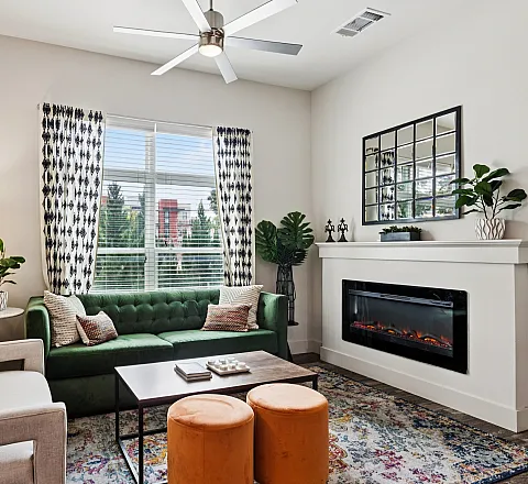 Chic living room with a modern green sofa, elegant armchair, and a cozy electric fireplace. Stylish decor elements include patterned curtains, vibrant artwork, and lush potted plants, creating a warm and inviting atmosphere.