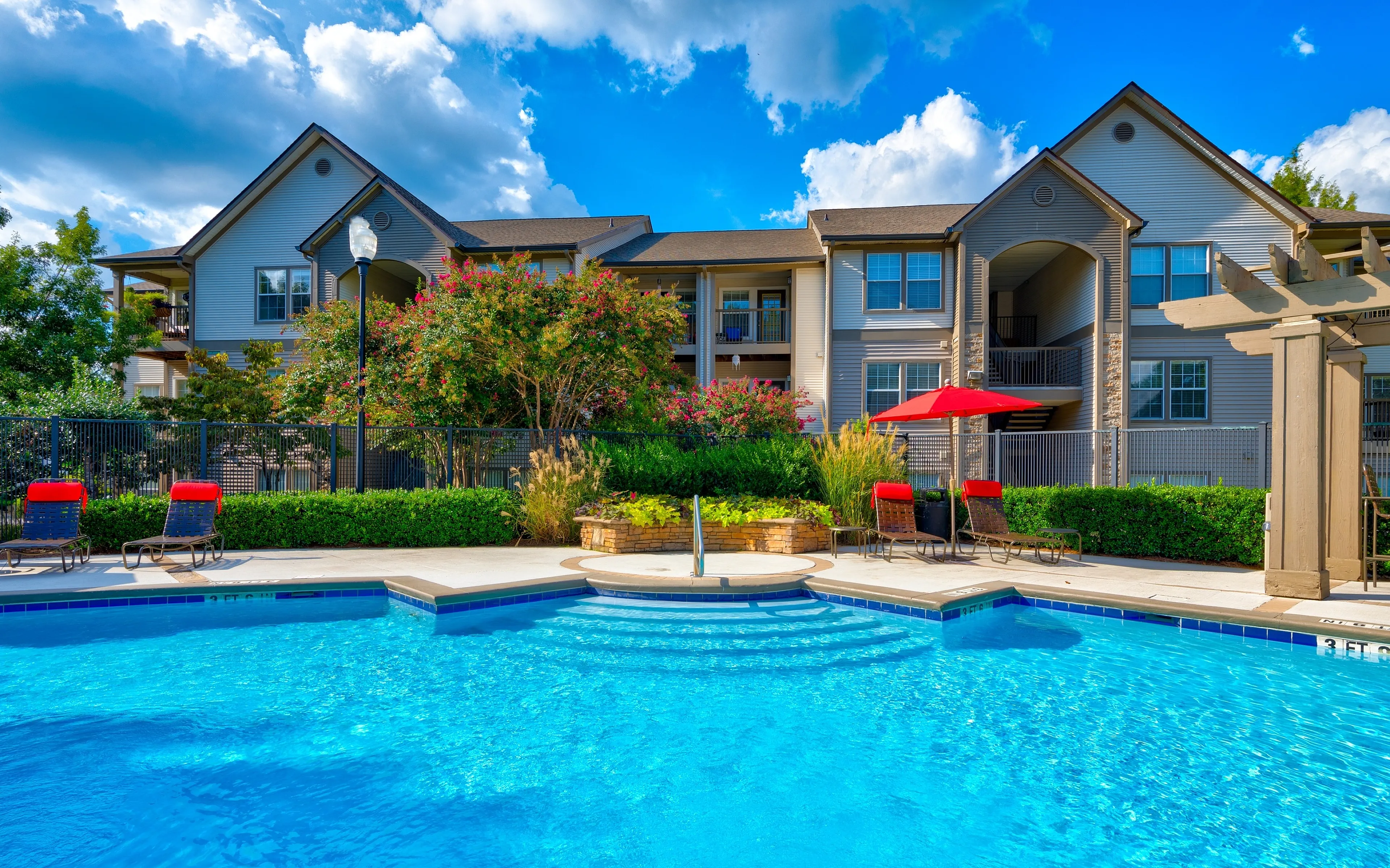 Serene apartment complex with a sparkling blue pool, lush landscaping, and cozy red lounge chairs under sunny skies.