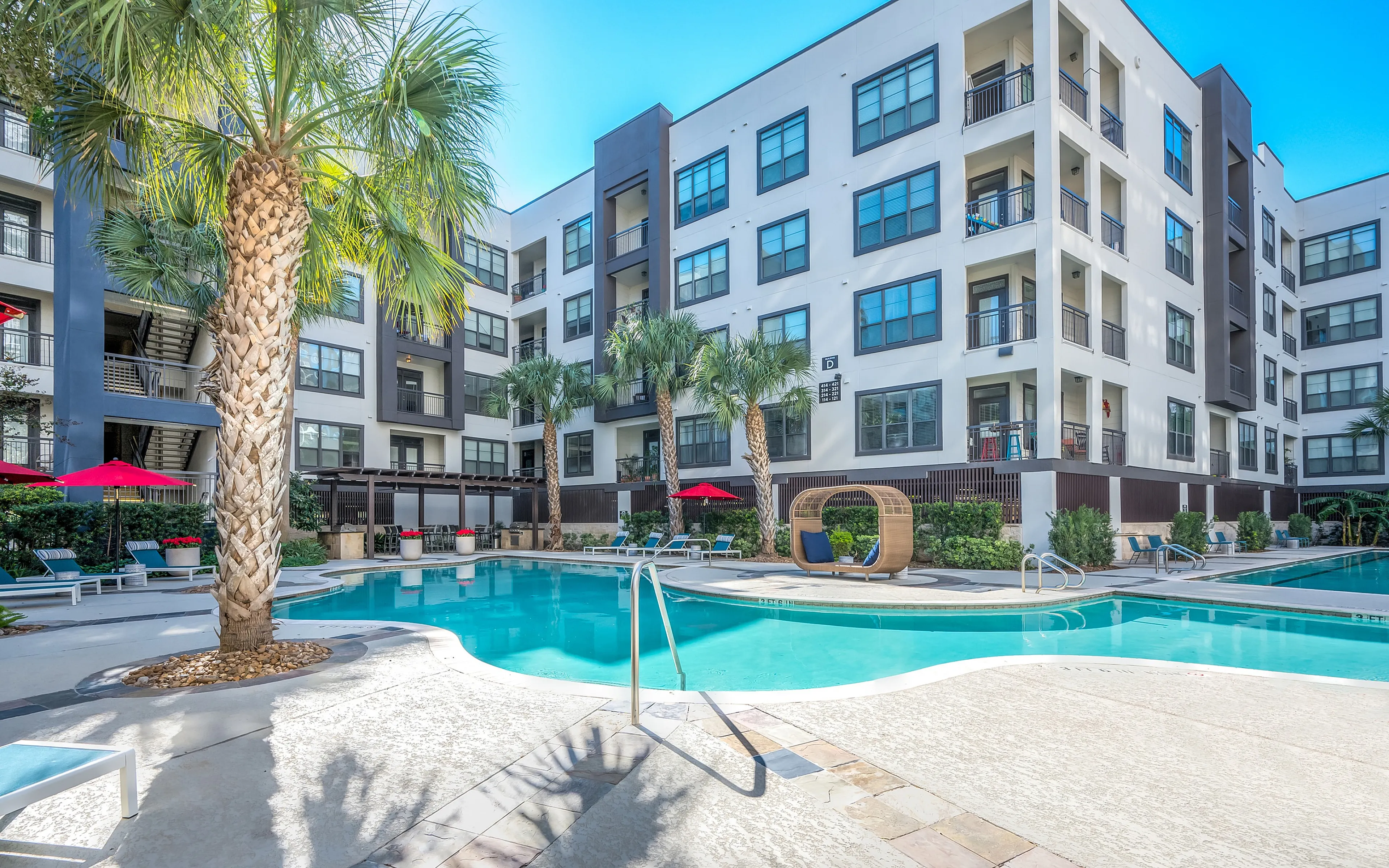 Luxurious apartment complex featuring a sparkling pool, elegant sun loungers, and lush palm trees, perfect for a tropical resort-style living experience.